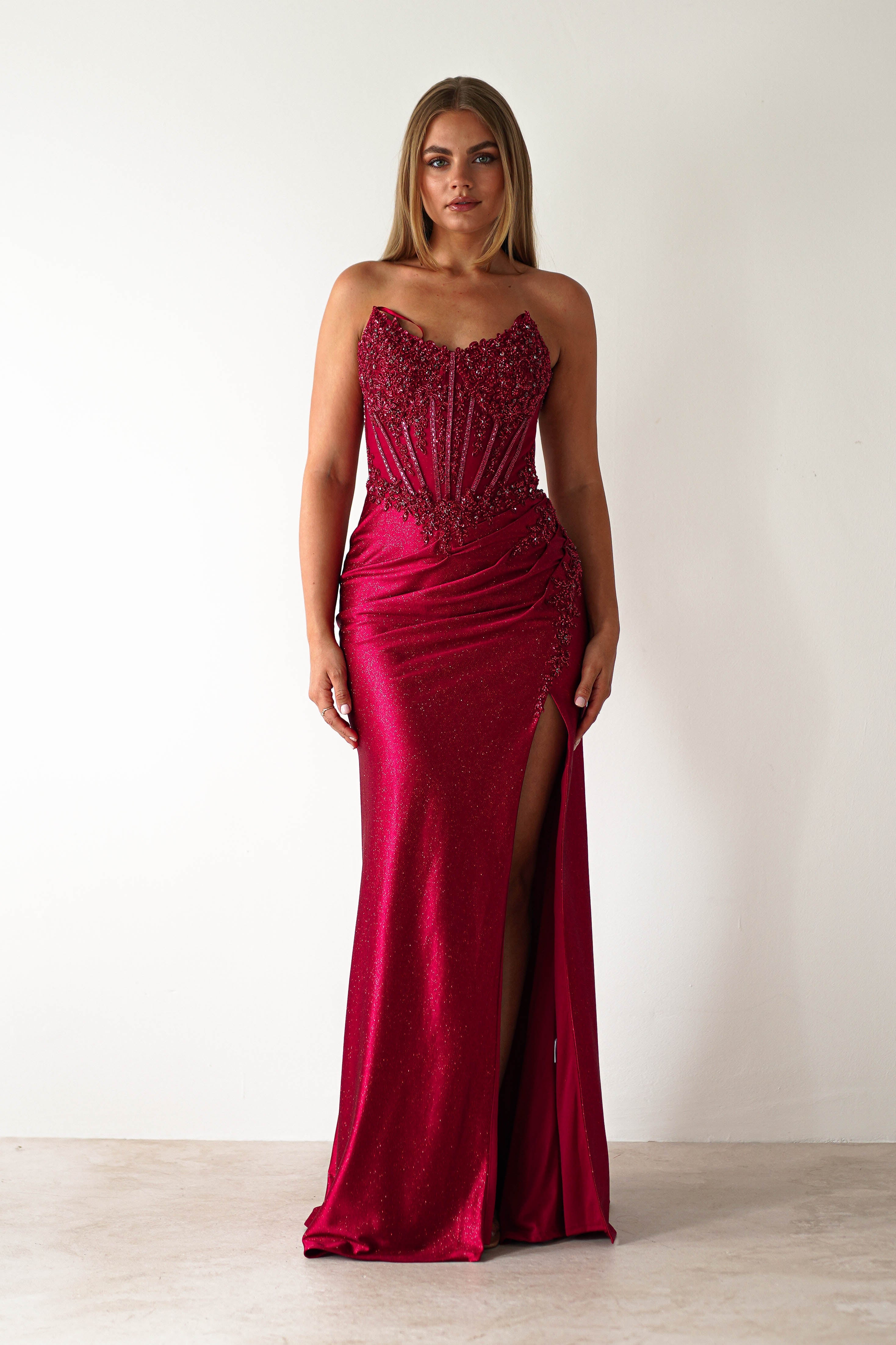 Woman wearing a deep red strapless bodycon gown with intricate beading, a structured bodice, and a thigh-high slit, standing against a white background, this dress is perfect for prom or debs.