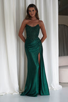 Woman wearing an emerald green strapless embellished gown with a corset bodice and thigh-high slit, standing in front of white curtains.