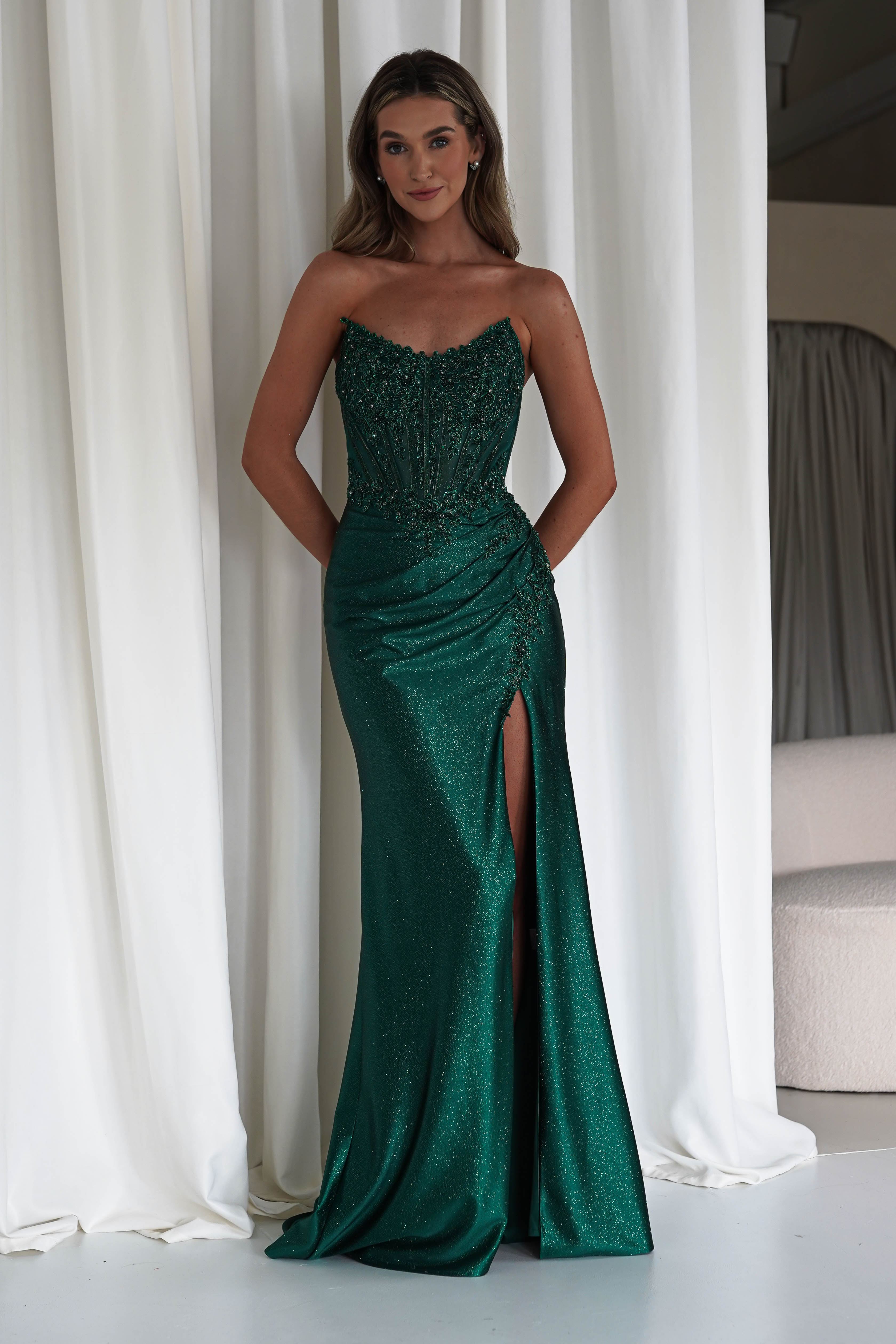 Woman wearing an emerald green strapless embellished gown with a corset bodice and thigh-high slit, standing in front of white curtains.