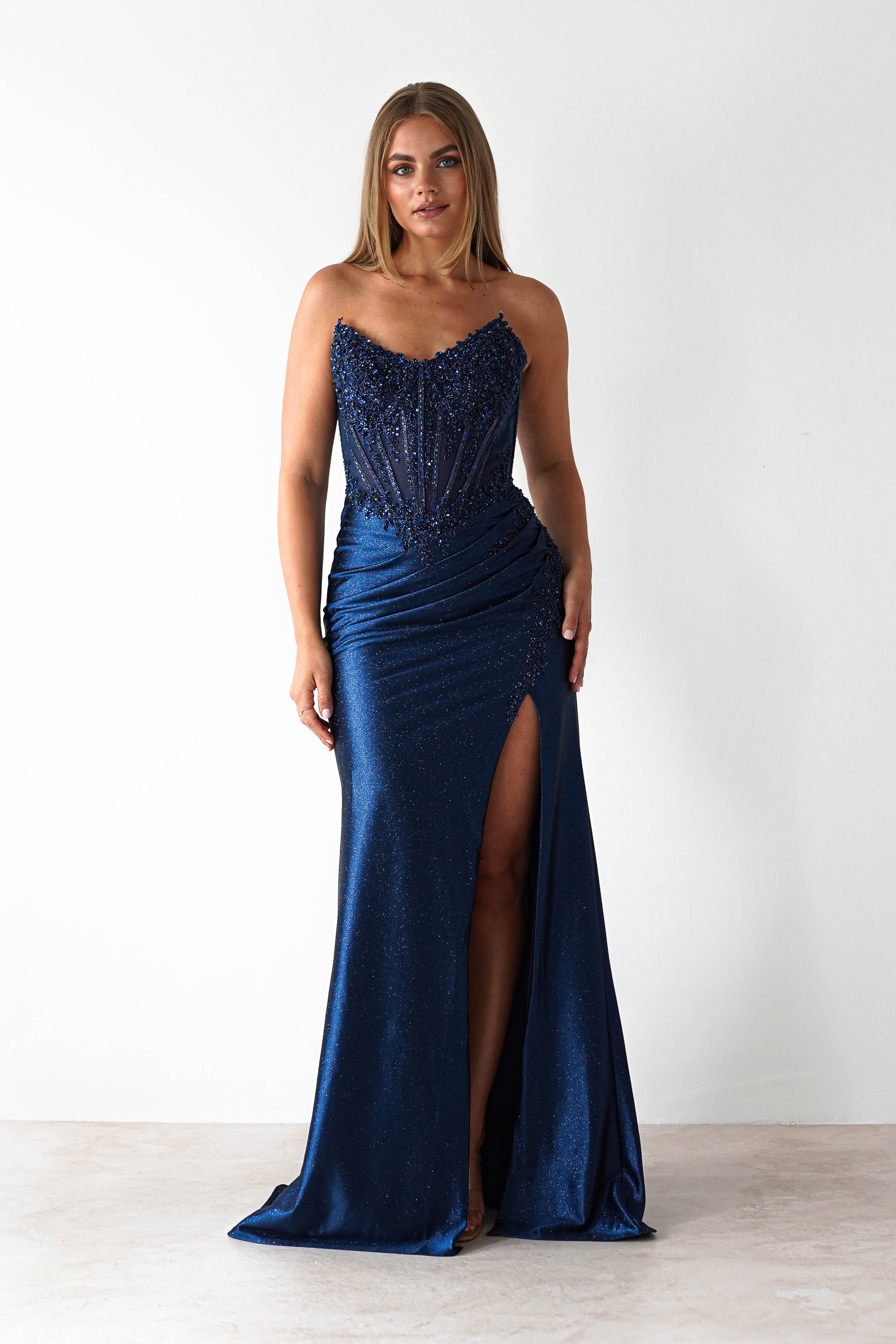Woman wearing a navy blue strapless bodycon prom gown with intricate beading, ruched detailing, and a thigh-high slit, posing against a white background.