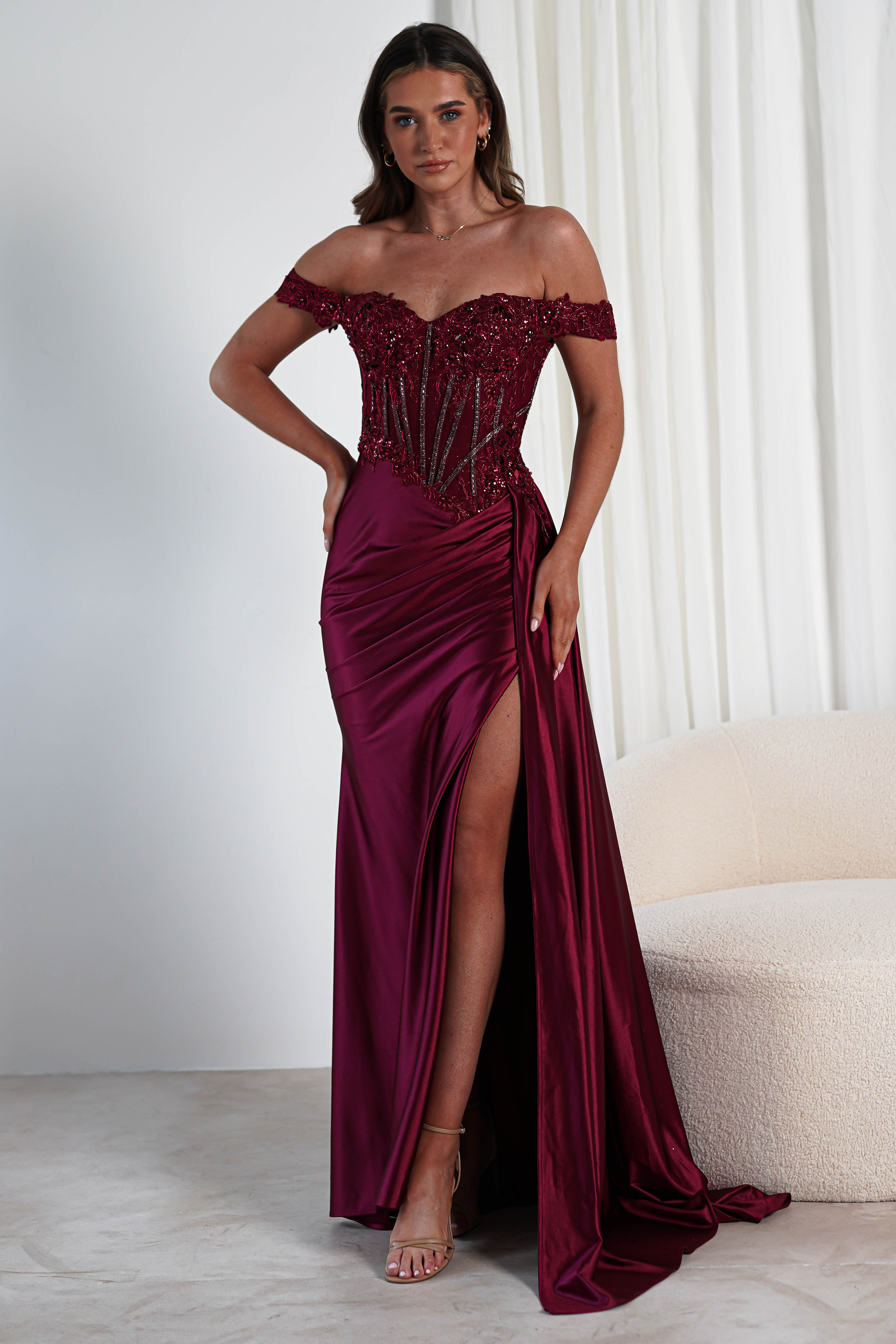 A woman stands wearing an off-the-shoulder, burgundy gown with intricate beadwork on the bodice, ideal for a formal occasion. The elegant gown features a thigh-high slit and a long, flowing skirt as she poses indoors next to a light-colored, modern chair.