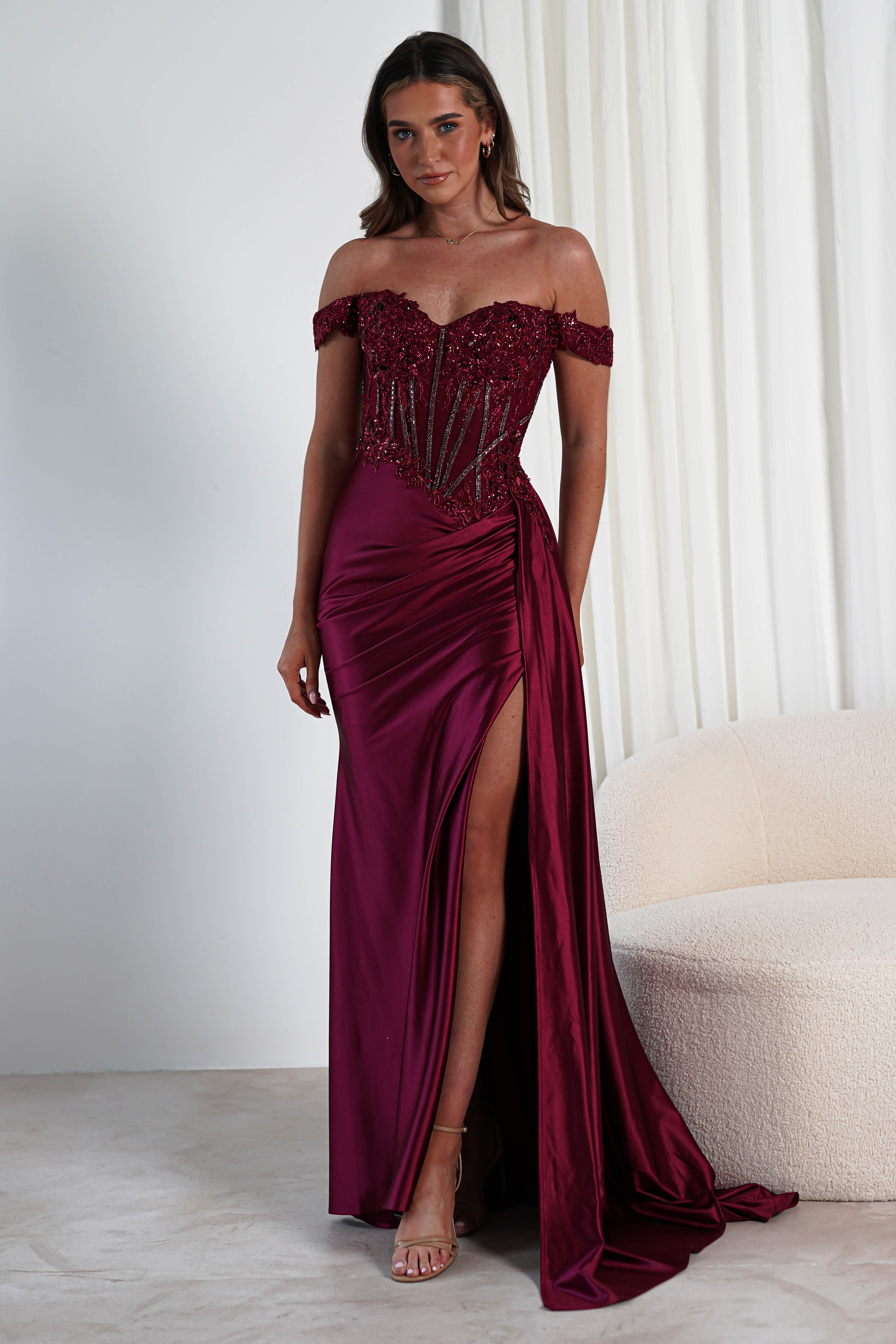 A woman in an elegant burgundy off-shoulder gown featuring a lace-embellished bodice and sequin details stands in a room with white curtains and a light beige curved sofa. Her long brown hair cascades down as she dons strappy heels, perfect for a formal occasion.