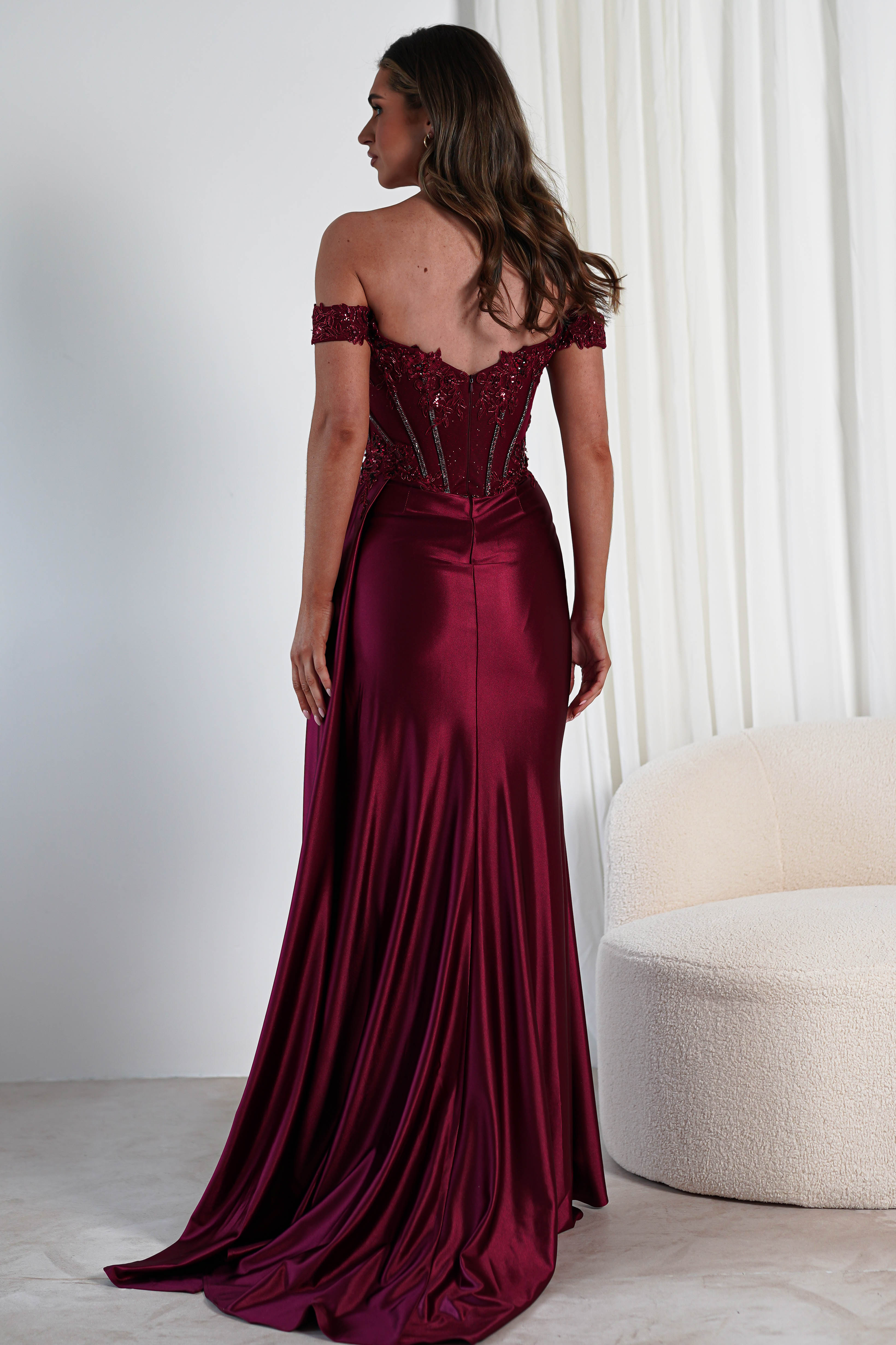 A woman faces away, wearing an elegant off-shoulder burgundy gown with a lace-embellished bodice and flowing train. Her long hair is styled naturally as she stands near a white fabric backdrop and a cream-colored curved chair, ready for the formal occasion.