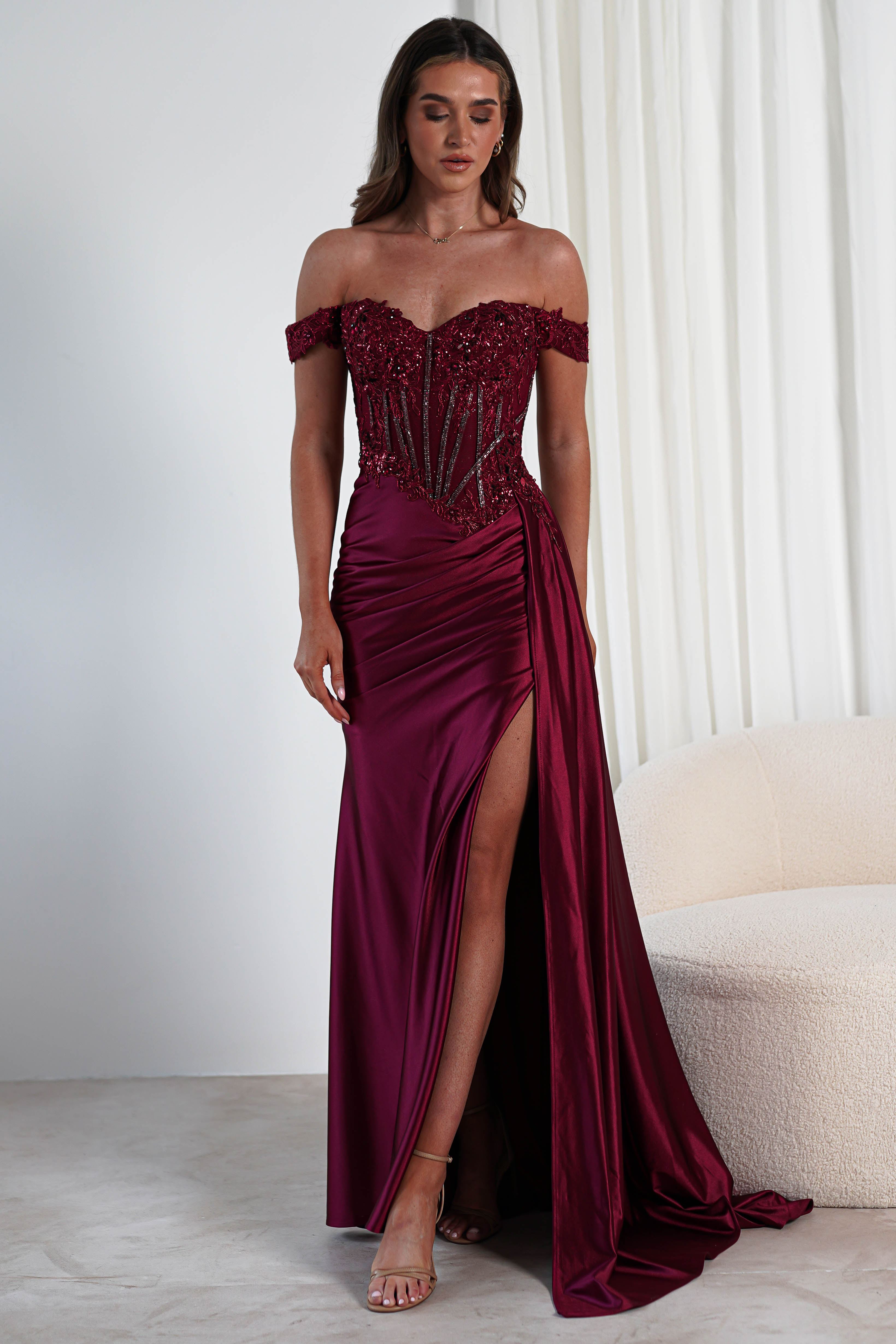 A woman in a burgundy off-shoulder evening gown with an elegant, lace-embellished bodice and a thigh-high slit stands in a room adorned with white curtains and a beige chair, embodying grace fit for any formal occasion.