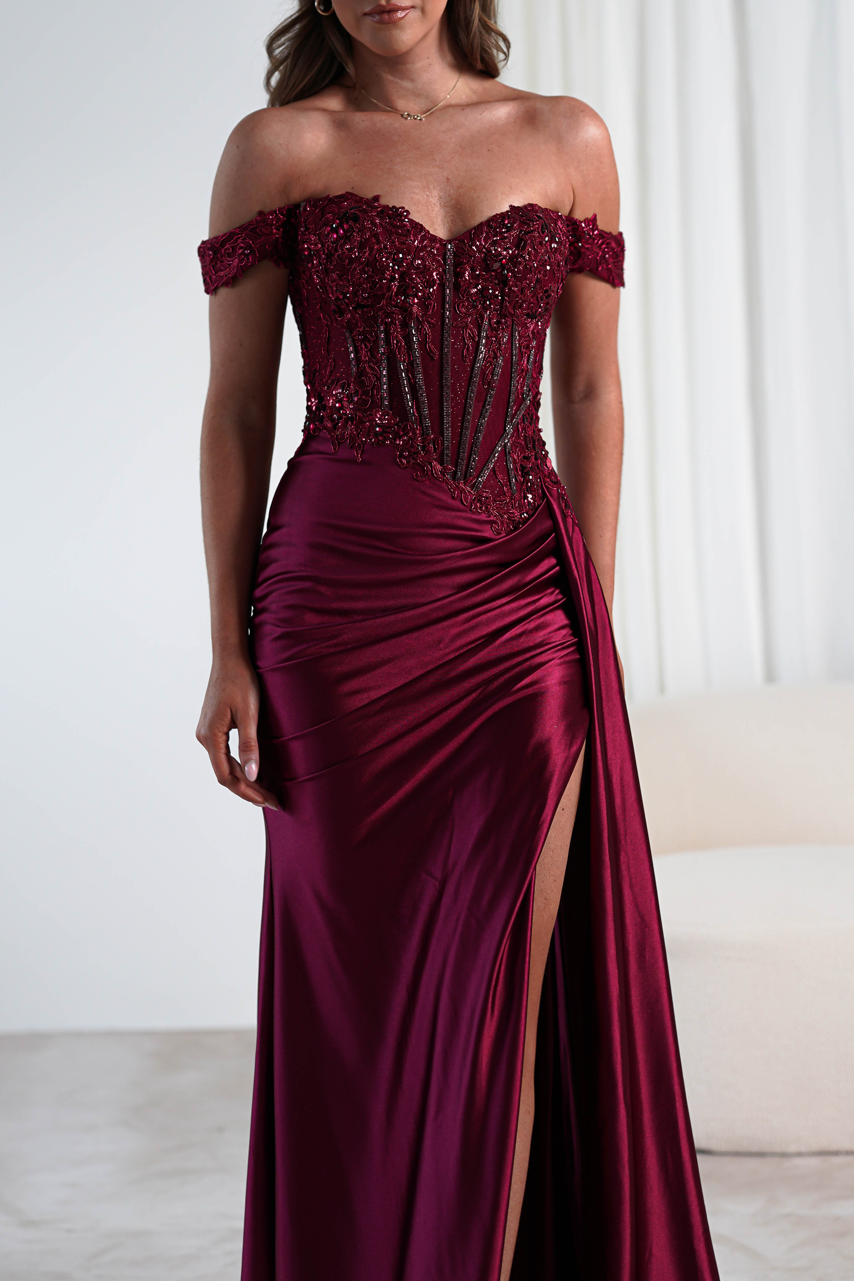 A woman stands in a softly lit room with white curtains, wearing an elegant burgundy gown perfect for a formal occasion. The off-the-shoulder dress features a lace-embellished bodice adorned with sequins and flows into a fitted silhouette with a daring thigh-high slit.