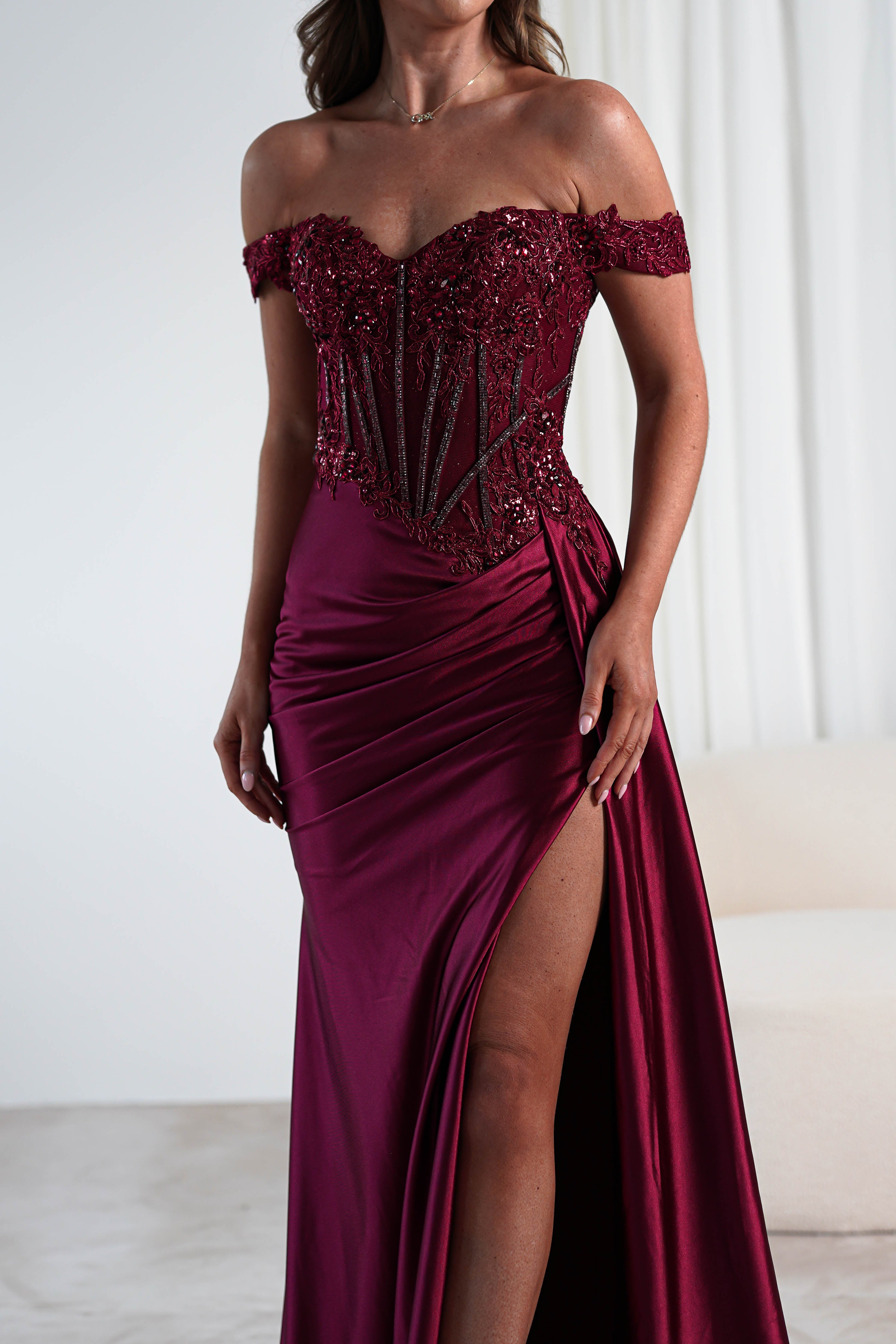 A person is wearing an off-the-shoulder, burgundy evening gown with a lace-embellished bodice and a high slit. The background is minimal, showcasing white curtains and a beige couch, perfect for a formal occasion.