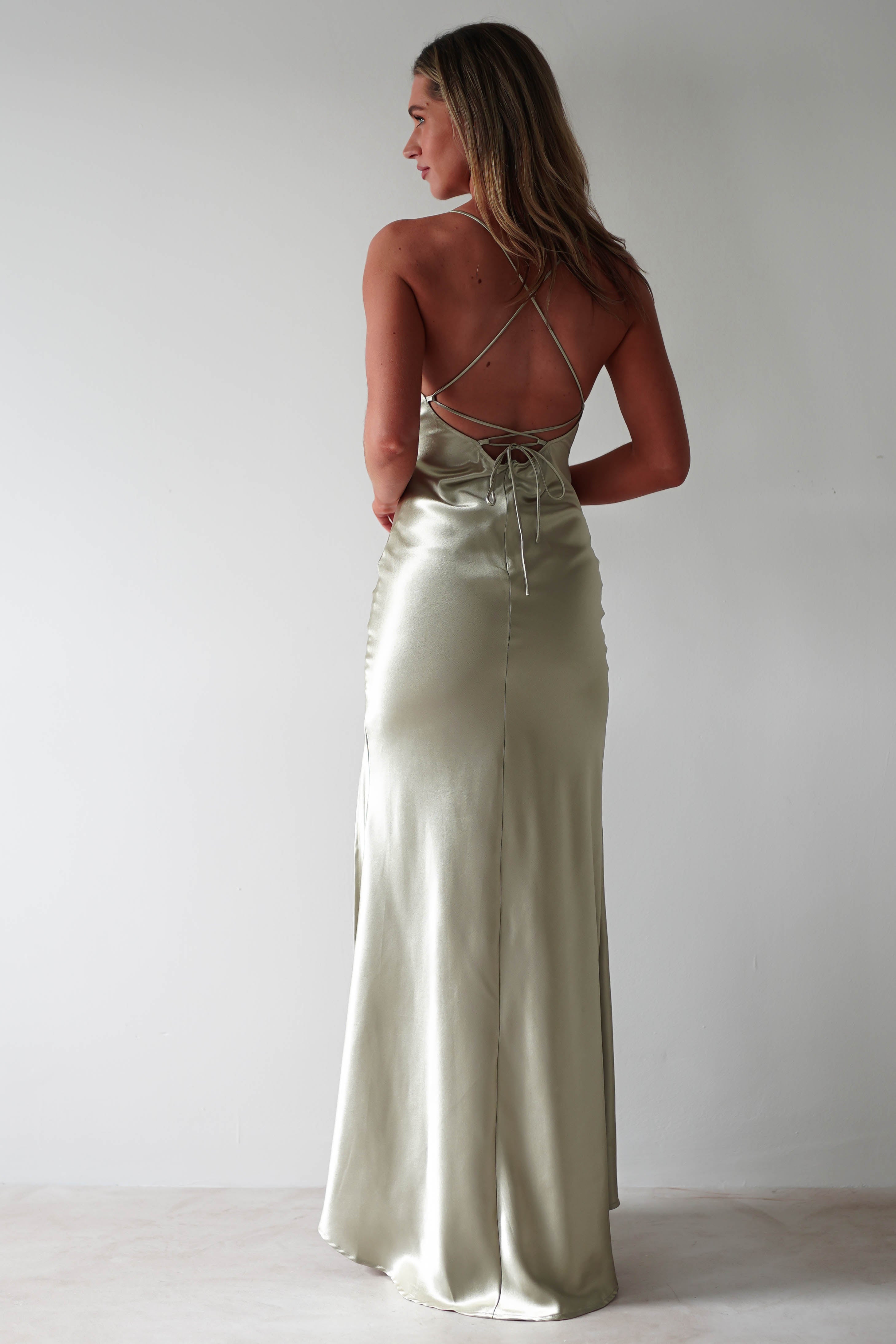 A woman stands facing away, wearing a light sage green satin slip dress with adjustable tie straps and a crisscross open back. The floor-length gown features a figure slimming, fitted silhouette and subtle sheen against a plain light background.