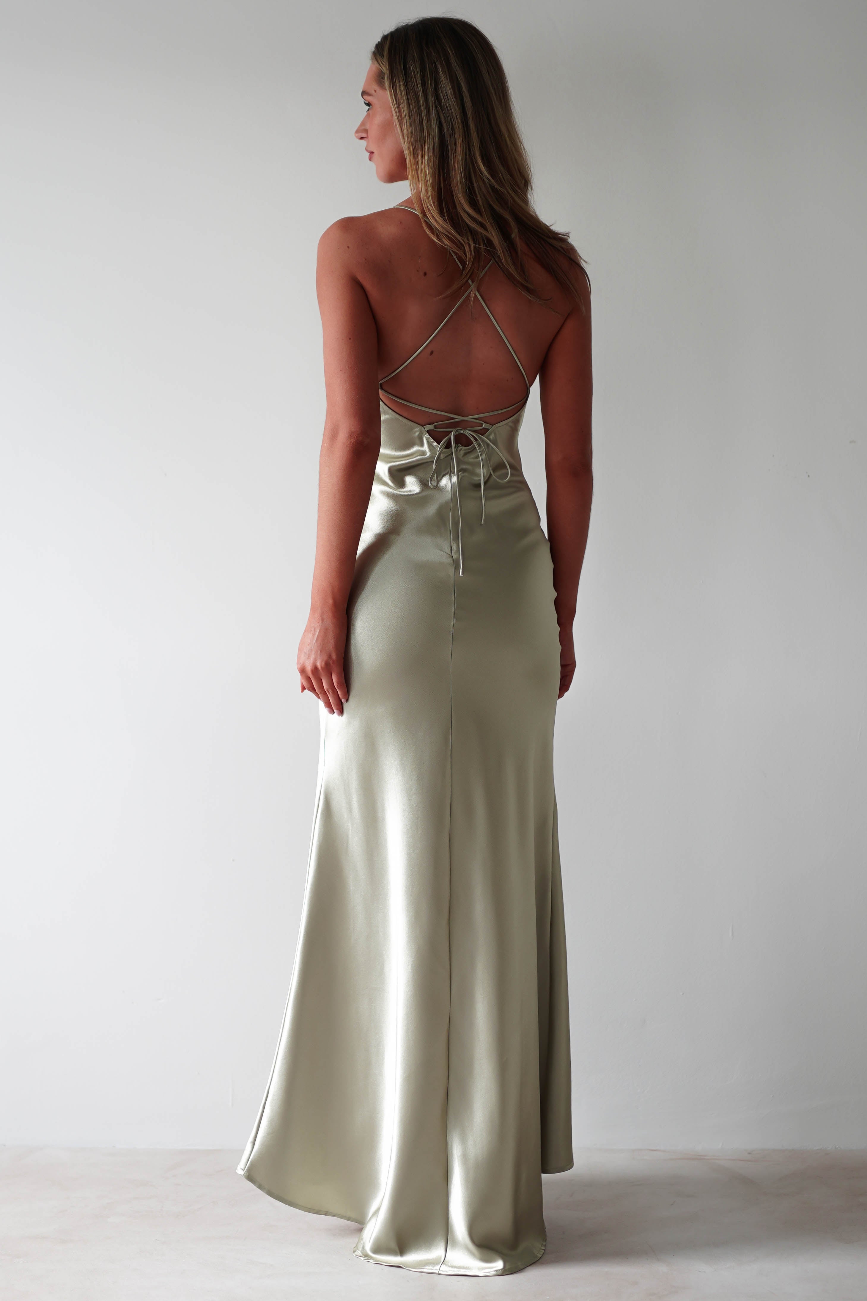 A woman stands facing away, wearing a long, light green satin slip dress with adjustable tie straps and an open back. The figure-slimming silhouette gently flares to the floor as she poses against a plain light background.
