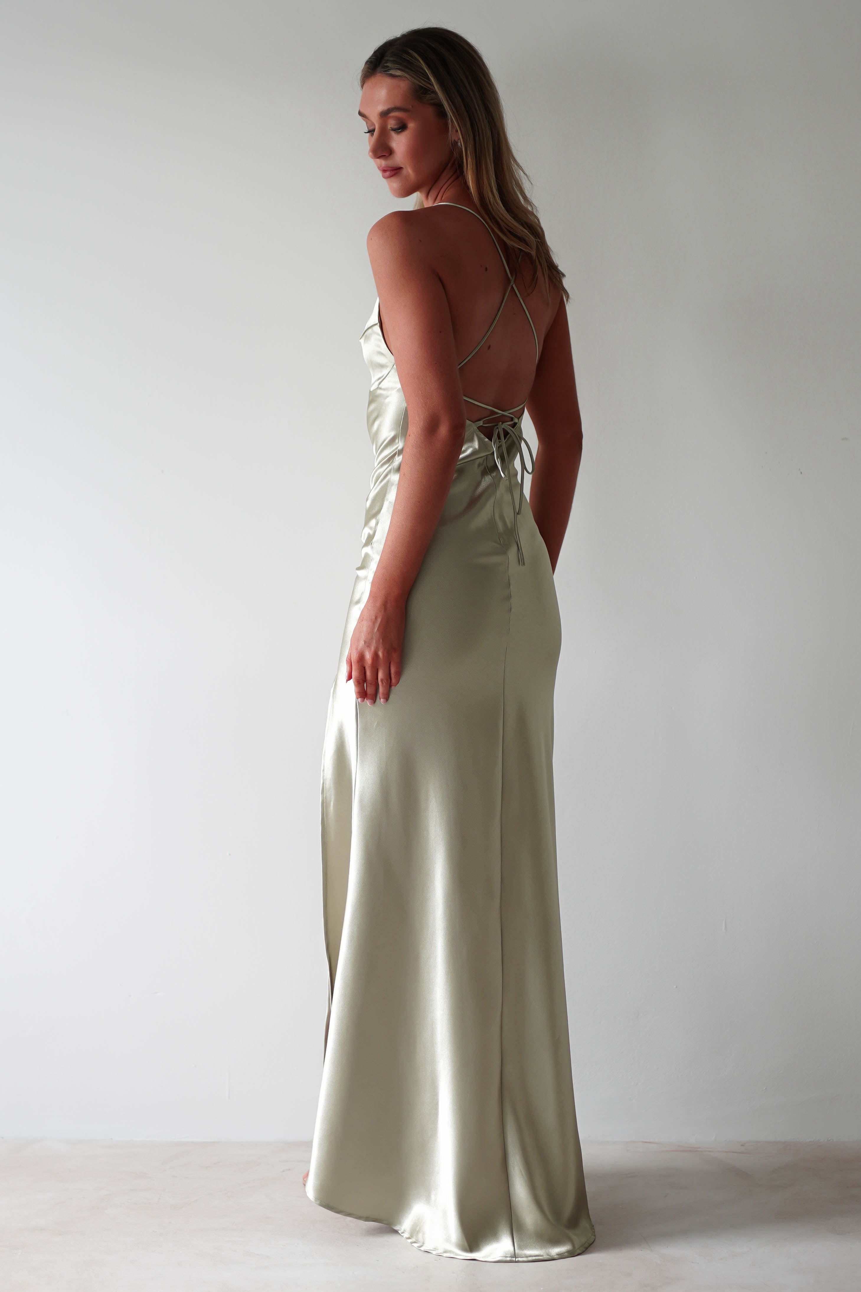 A woman in a light, silky, floor-length slip dress with figure-slimming lines and adjustable tie straps stands against a plain light wall, looking over her shoulder.