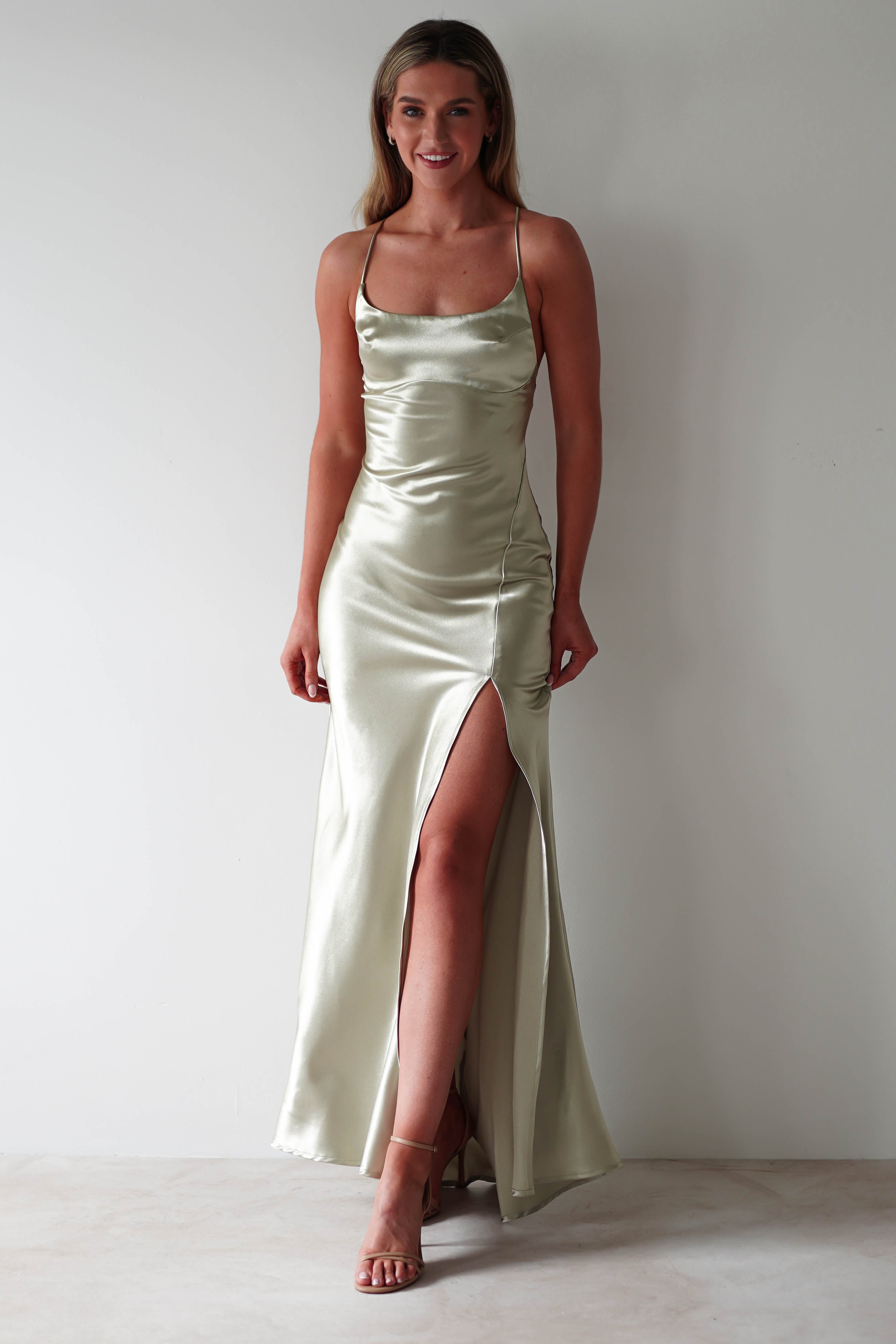 A woman stands against a white wall, smiling in a light green satin slip dress with adjustable tie straps and a thigh-high slit. The figure slimming design pairs perfectly with her nude heels, while her hair falls loosely around her shoulders.