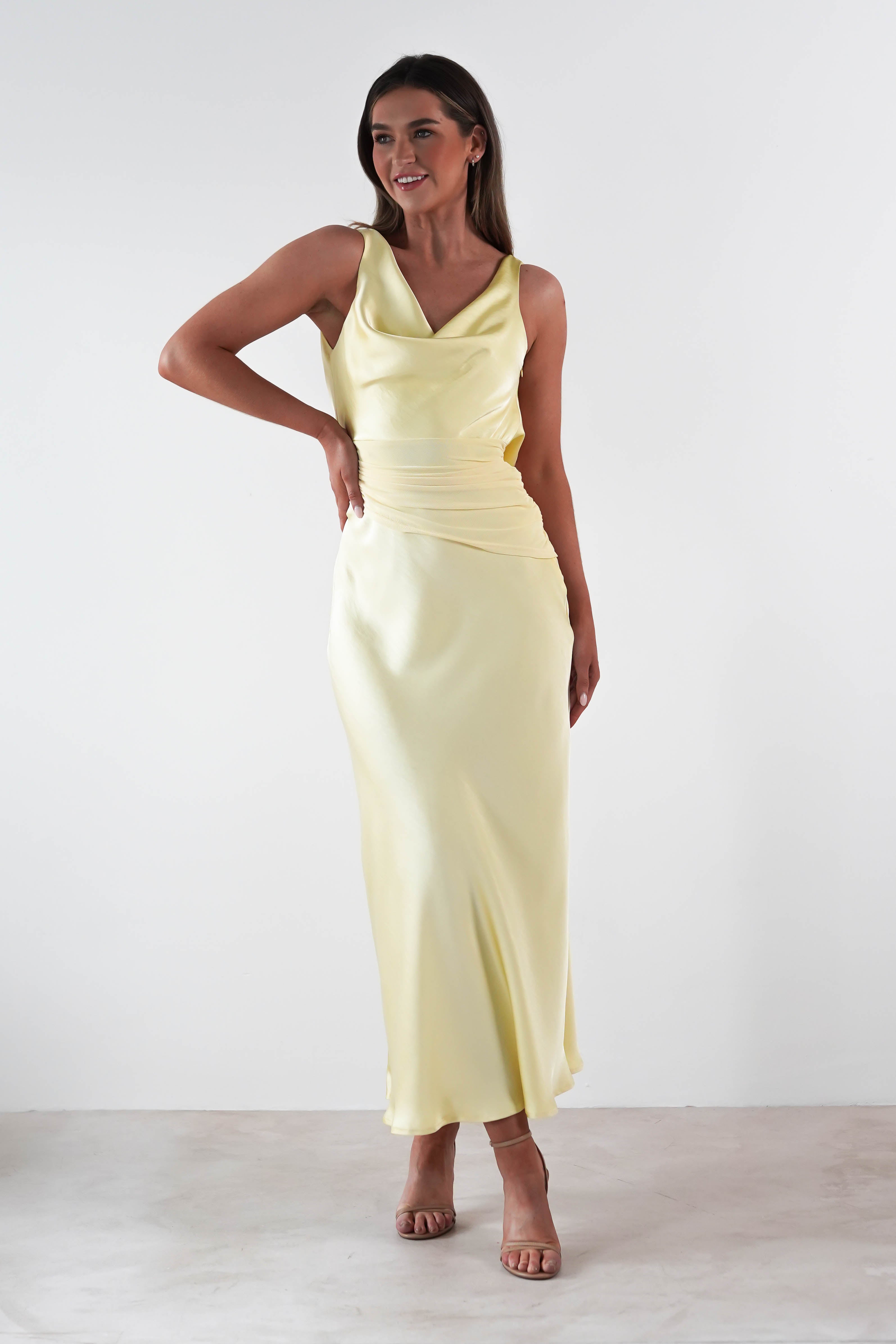 A woman in a sleeveless, floor-length satin maxi dress stands posing with one hand on her hip. She is wearing high heels and the background is plain white.