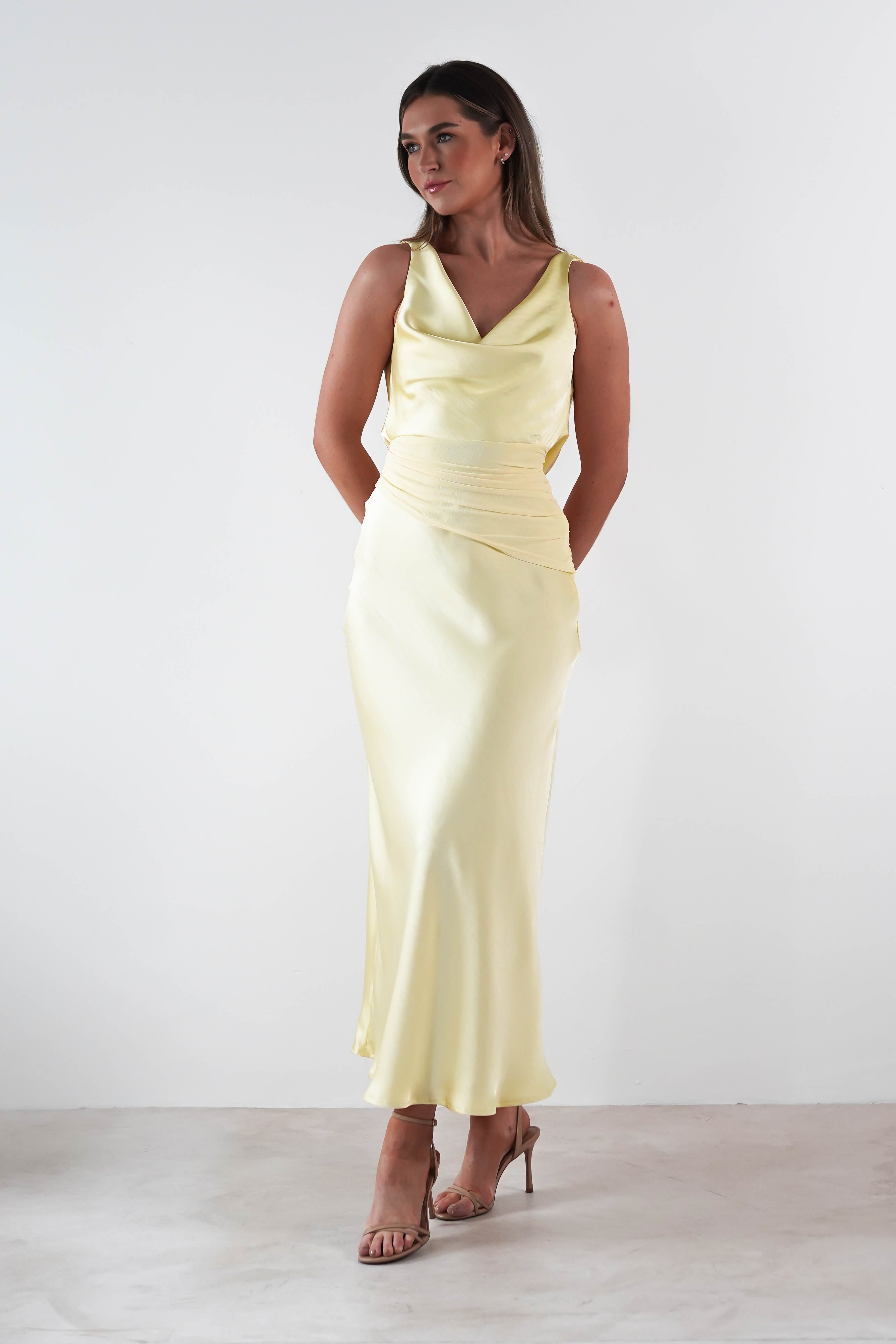 A woman stands against a plain white background, wearing a stunning satin maxi dress in pale yellow, featuring an elegant draped neckline. She pairs the wedding guest dress with strappy high heels, her hands gracefully tucked behind her back.