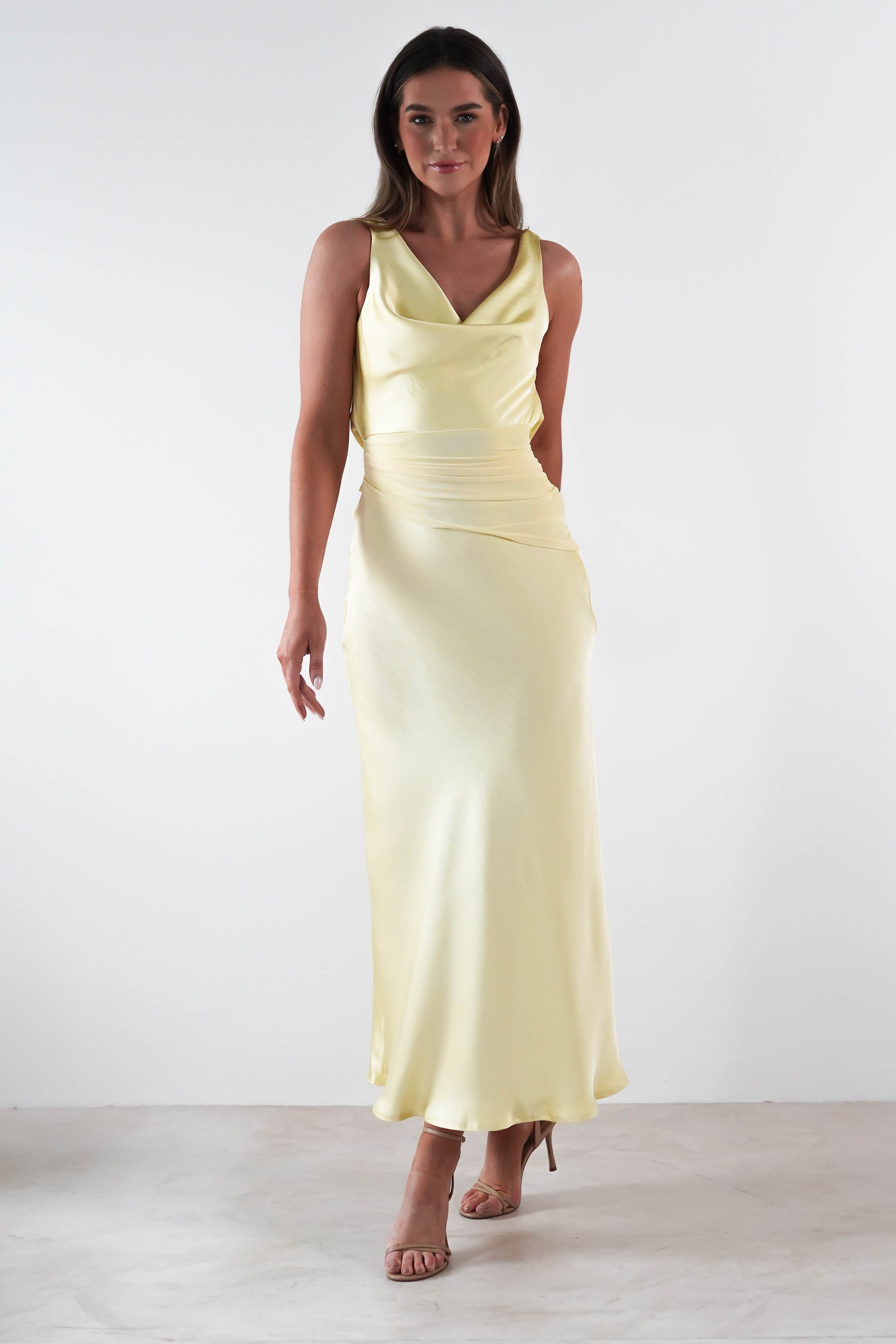 A woman graces the scene in a satin maxi dress, its long, light-yellow fabric cascading elegantly with a cowl neckline. Standing against a plain white background, she showcases open-toed heels and long hair, embodying the perfect wedding guest dress aesthetic.