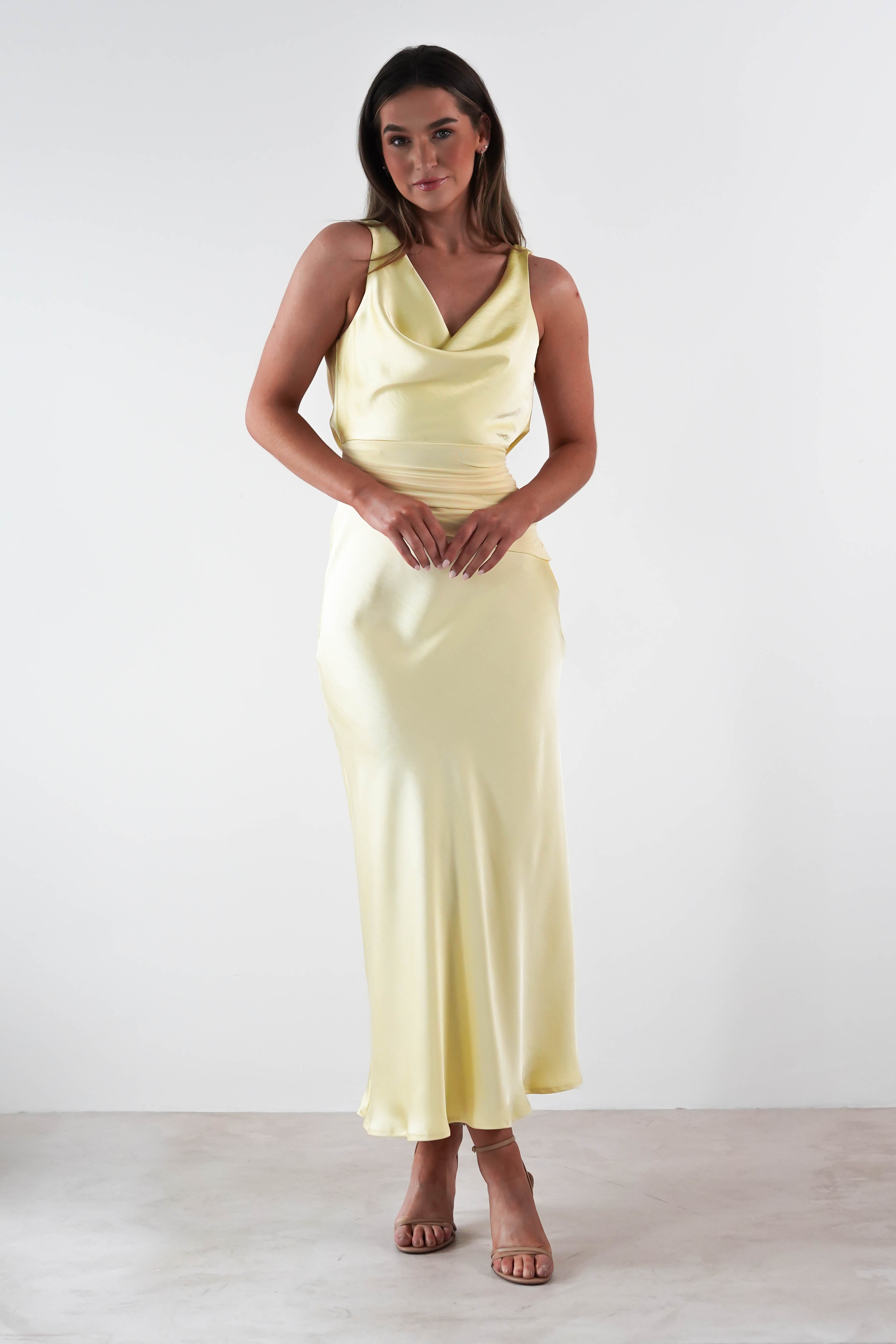A person stands against a plain white background wearing a light yellow satin maxi dress. They have long hair and are wearing open-toed heels. Their hands are gently clasped in front, embodying elegance perfect for a wedding guest dress.