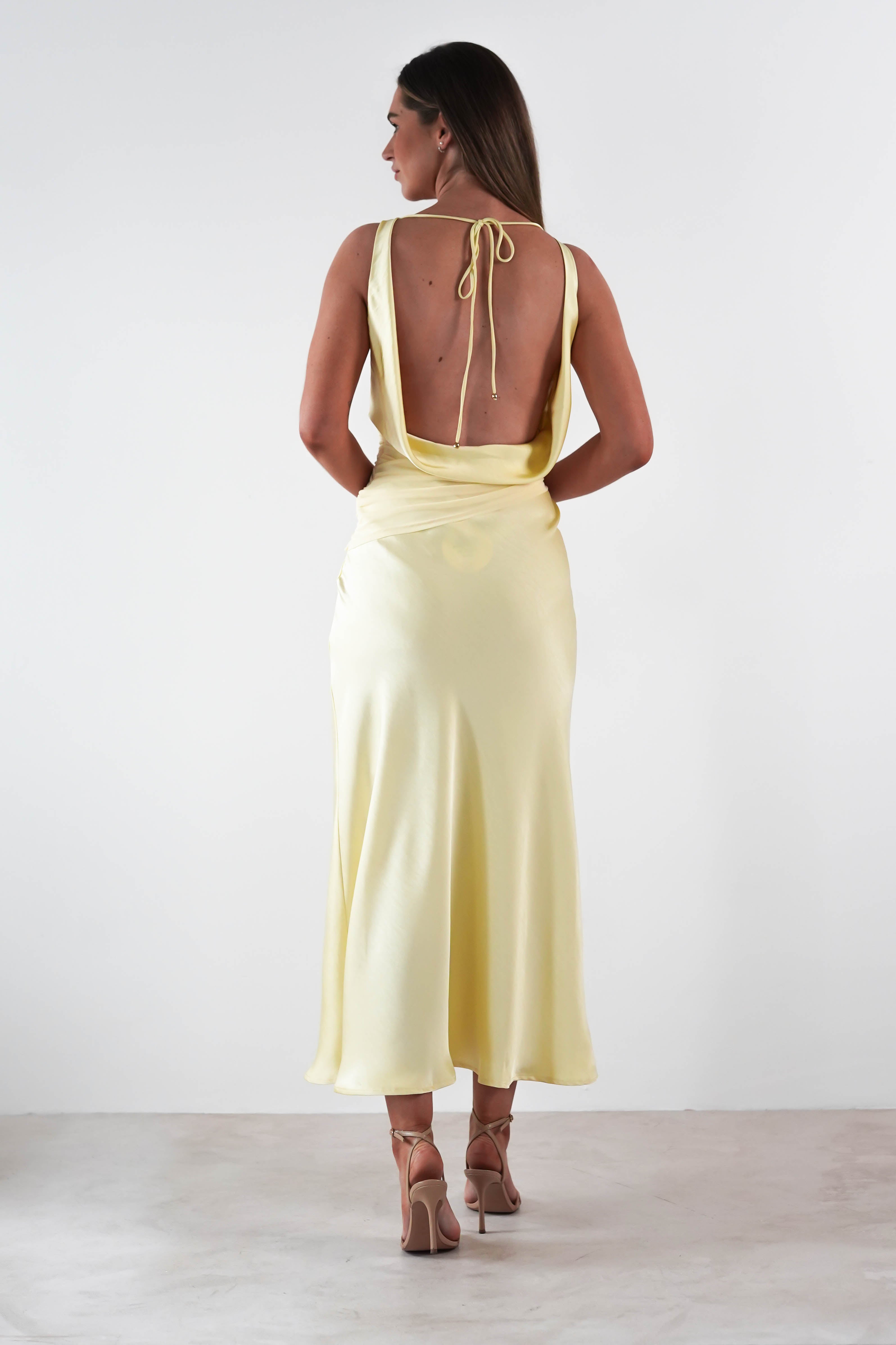 A woman stands with her back to the camera, wearing a light yellow satin maxi dress with tied straps and strappy heels. The dress features a long, flowing skirt, perfect for a wedding guest. Her long hair is down against the plain white background.