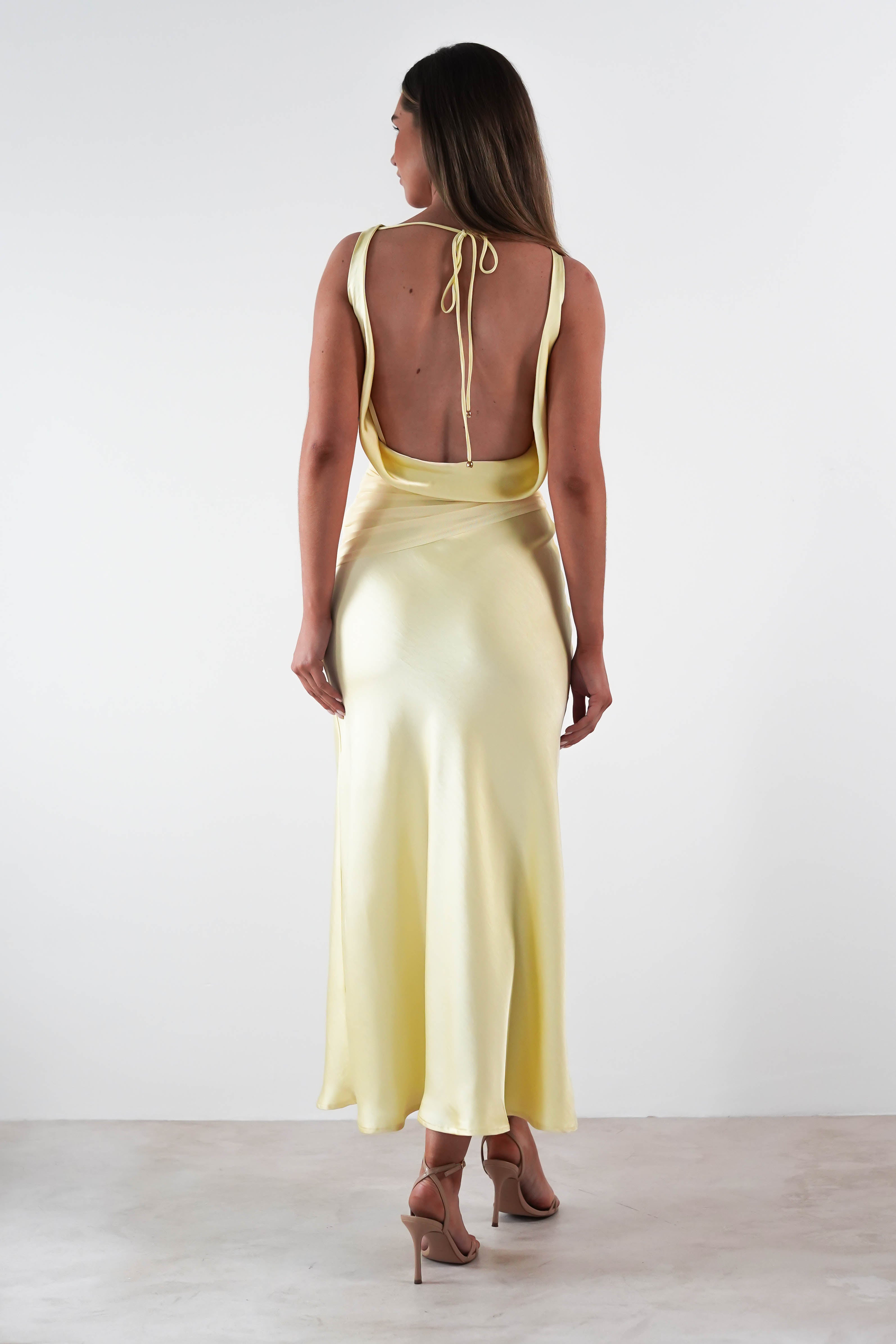 A woman stands with her back to the camera, wearing a stunning sleeveless yellow satin maxi dress featuring an open back and tie detail. Her high heels complete the elegant look against the plain white wall, perfect as a chic wedding guest dress.