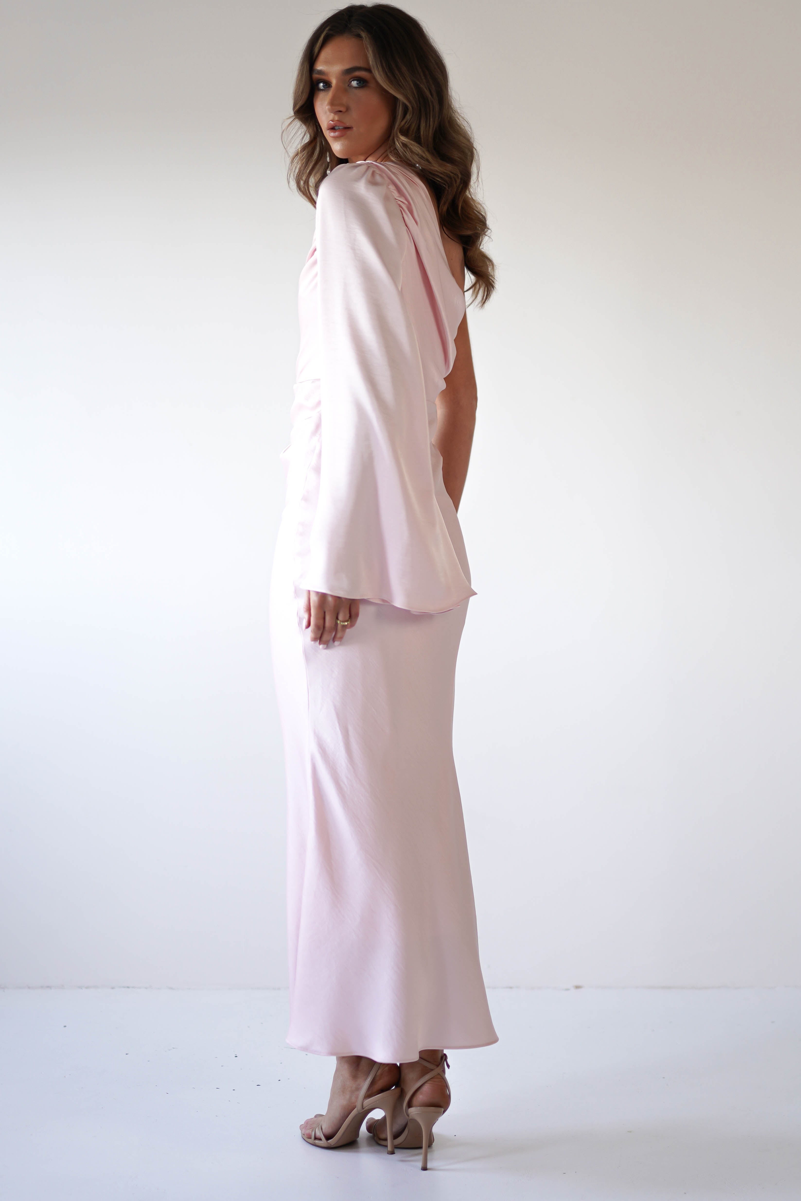A woman in a pale pink, floor-length dress with an asymmetrical neckline and long, draped sleeves stands in profile against a plain white background, wearing high-heeled sandals and looking over her shoulder.
