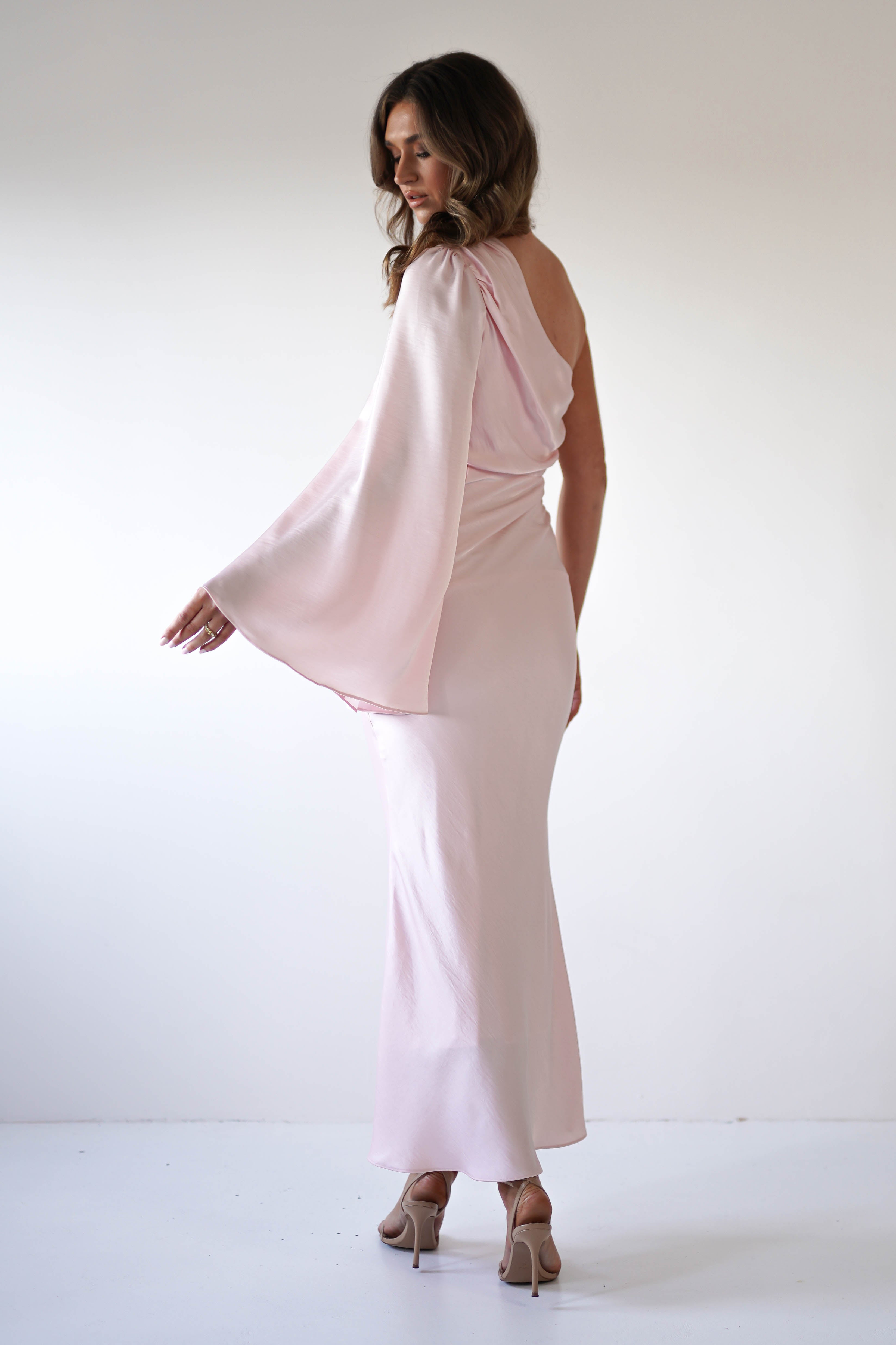 A woman in a pale pink, floor-length gown of silky fabric with an asymmetrical neckline and flowing cape sleeve stands in heels against a plain white background, looking over her shoulder.