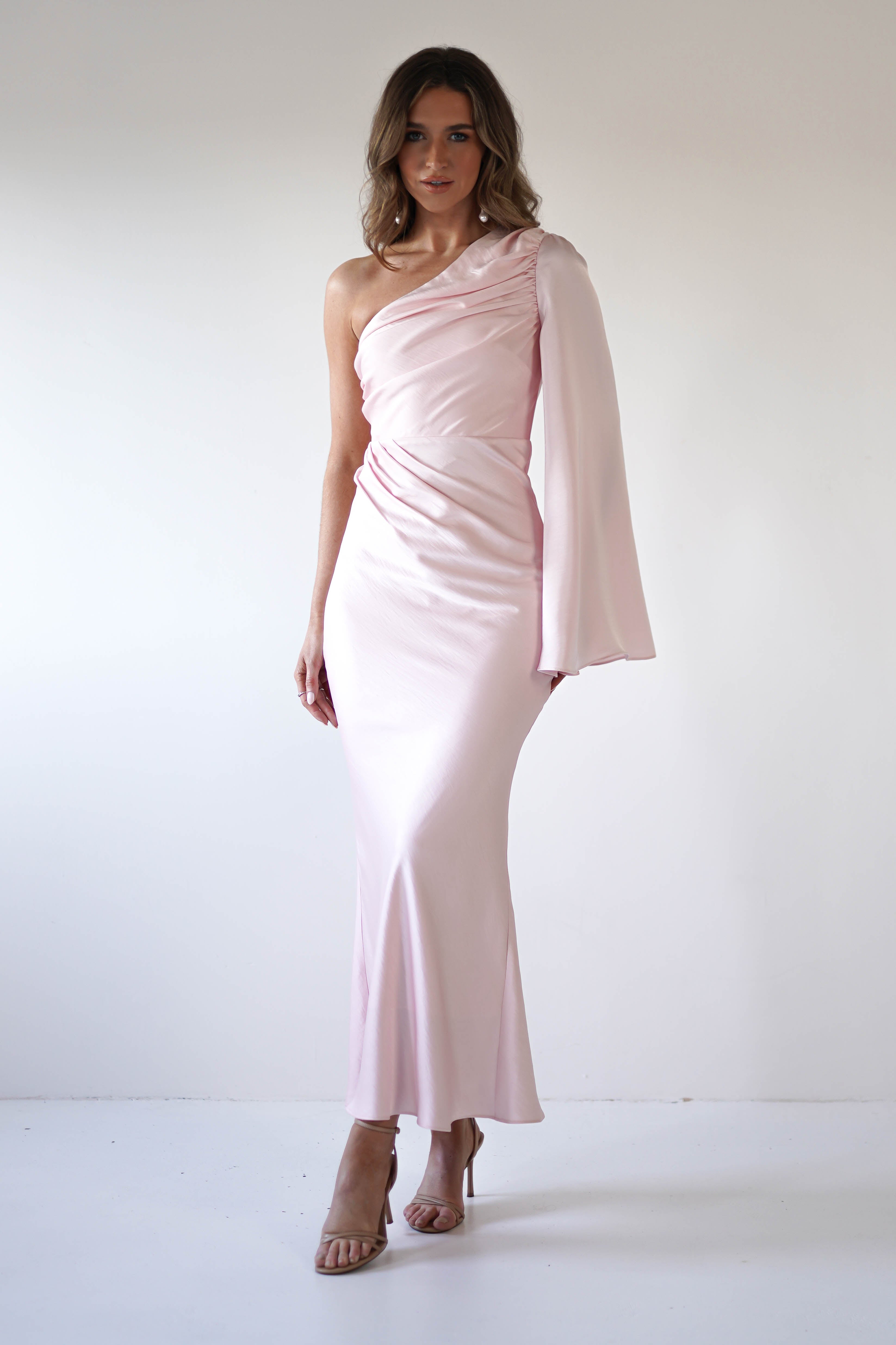 A woman stands against a plain white background wearing a light pink, silky fabric one-shoulder, floor-length dress with gathered detailing, a draped sleeve, an asymmetrical neckline, and nude heels.