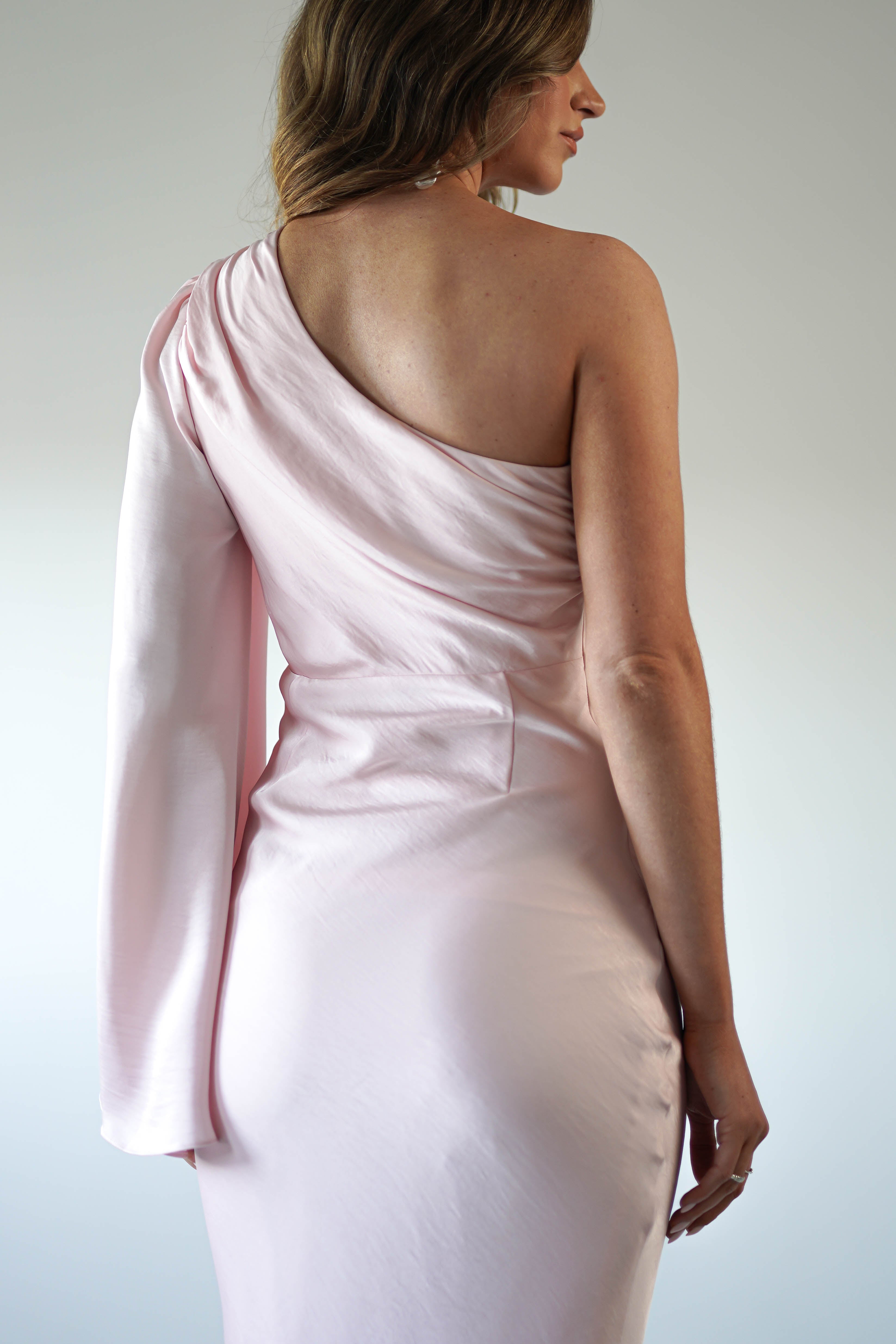 A woman in a light pink, one-shoulder dress with an asymmetrical neckline and gathered detailing stands facing away, highlighting the silky fabric and elegant design against a neutral background.