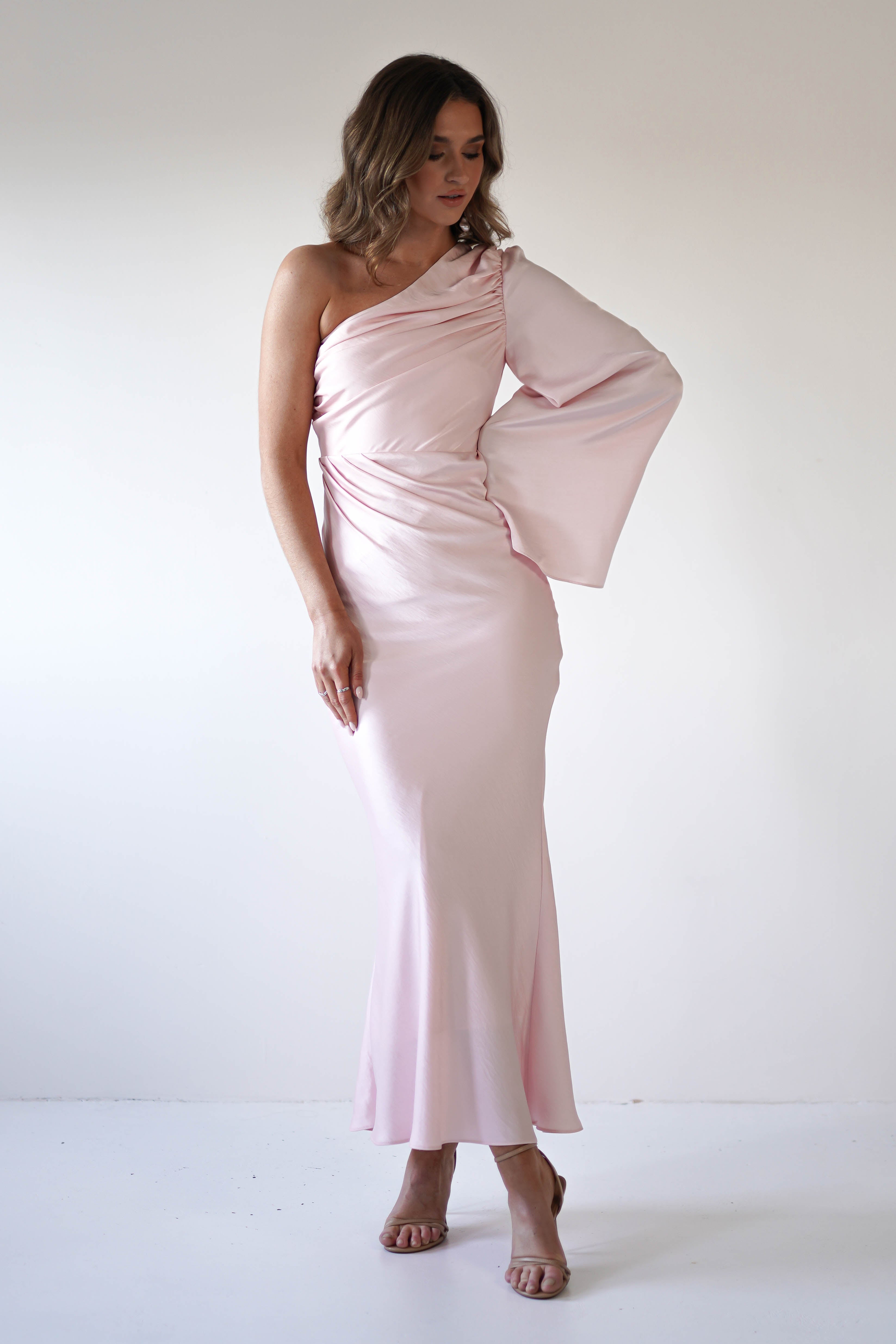 A woman in a pale pink, silky fabric gown with an asymmetrical neckline and flared sleeve stands against a plain white background, looking down with one hand resting on her hip.
