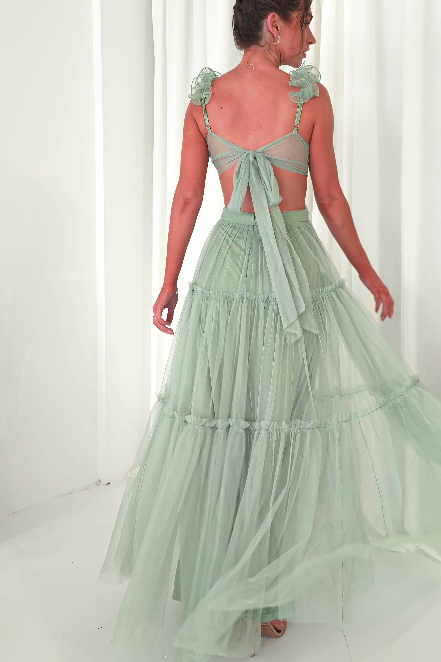 In a softly lit room with white curtains, a woman poses with her back turned, draped in a light green tulle maxi dress featuring ruffled straps and a tied back. The dresss elegant flow makes it perfect for any wedding guest looking to make an impression.