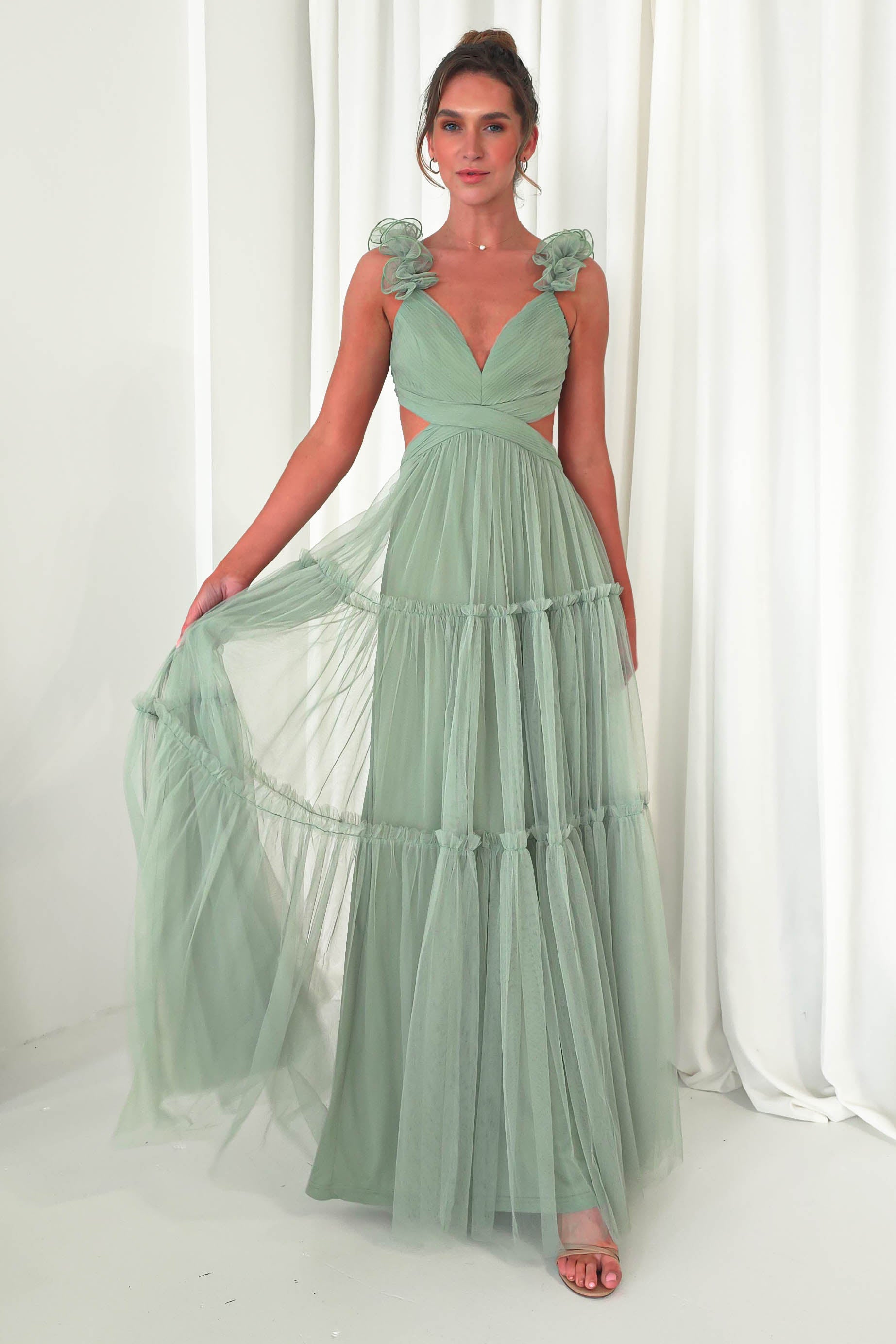 A woman poses gracefully in a flowing pastel green tulle maxi dress, featuring ruffle details and stylish cutouts at the waist. The gown, perfect for a wedding guest, boasts a deep V-neckline and tiered layers against a backdrop of white curtains.