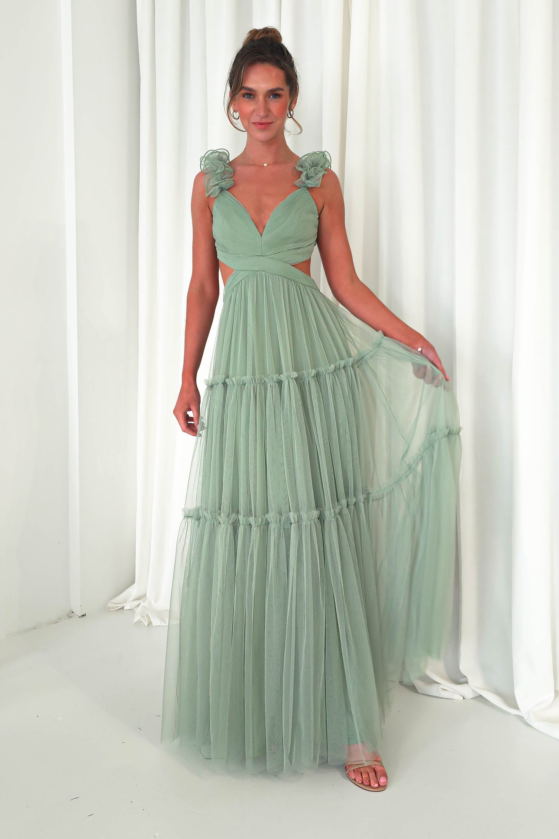 A person stands in front of white curtains, wearing a flowing mint green tulle maxi dress with a deep V-neckline and ruffled shoulder accents. Their hair is styled up, and they hold a piece of the dress to the side, bare feet visible—perfect attire for a wedding guest.