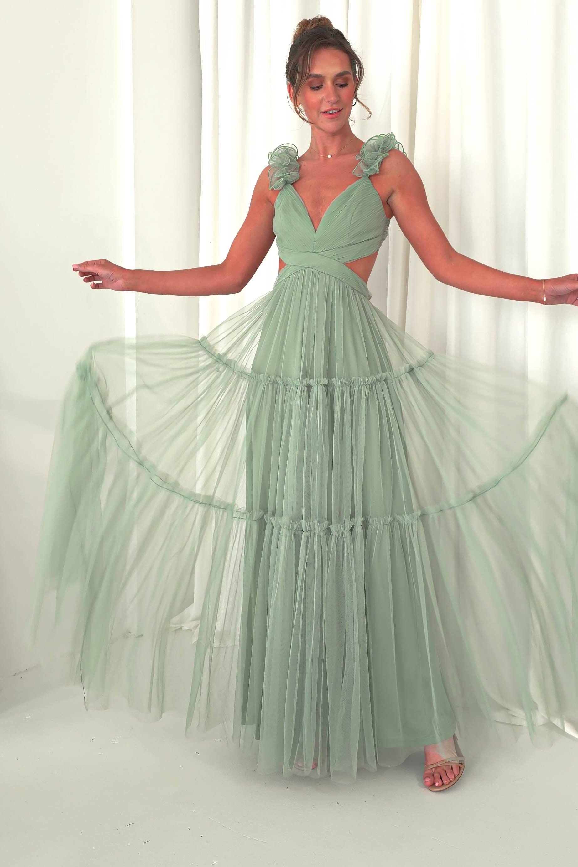 A woman poses gracefully in a flowing, light green tulle maxi dress. The dress, ideal for a wedding guest, boasts floral details on the shoulder straps and a deep V neckline. She stands in front of a white curtain, gently holding the dresss sides to display its layers.