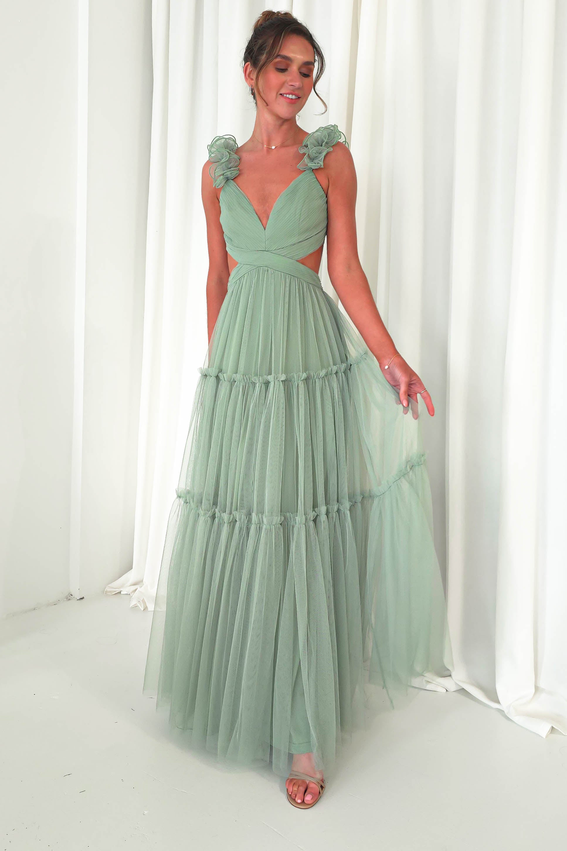 A woman poses in a flowing mint green tulle maxi dress with ruffled shoulder straps and a V-neckline. The gown, perfect for a wedding guest, features layered tulle and cut-out sides. She stands in front of a white curtain, smiling and looking to the side.