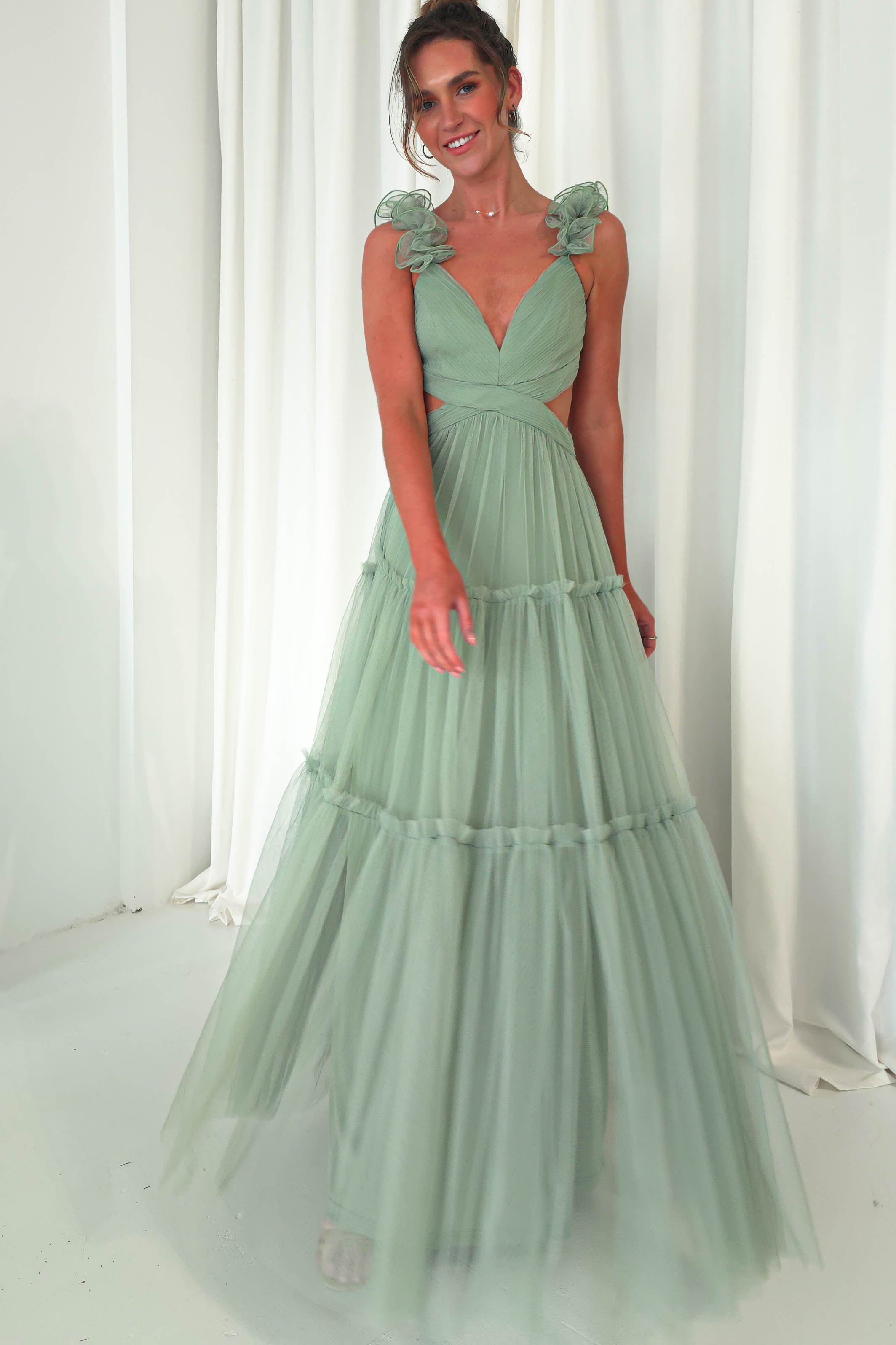 A person wearing a long, flowing tulle maxi dress with ruffled shoulder straps stands against a white curtain backdrop. Smiling as a joyful wedding guest, one hand is by their side while the other is slightly raised, showcasing the elegance of their attire.