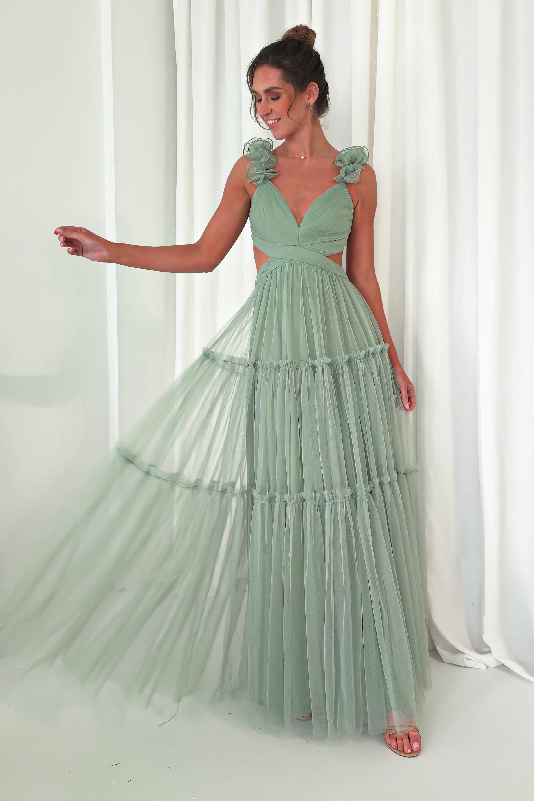A woman in a sage green, sleeveless tulle maxi dress with ruffled straps poses against a white curtain backdrop. Smiling, she elegantly holds the gown to one side, showcasing its chic cut-out waist—perfect for a fashionable wedding guest look.