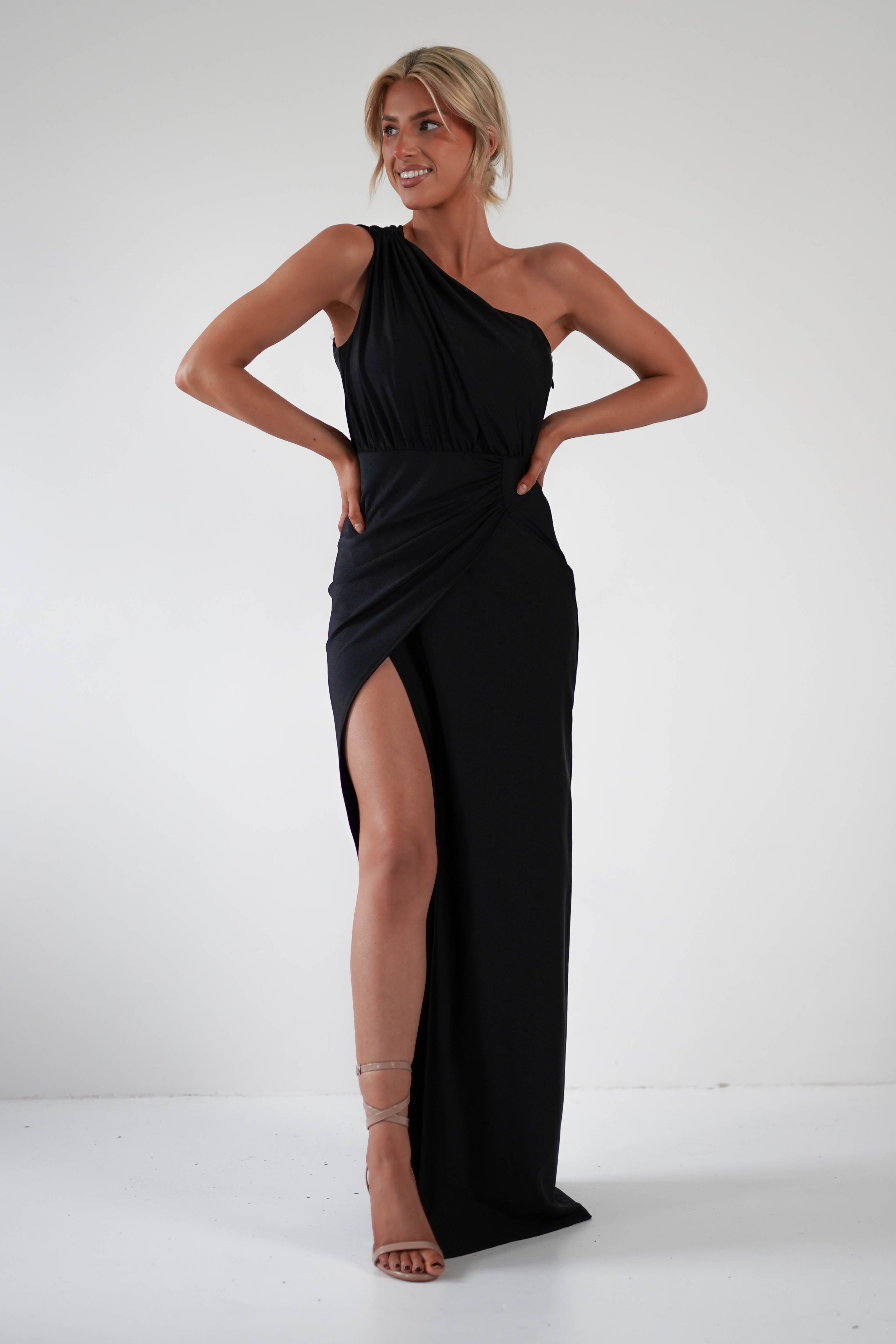 A person poses confidently in a sleek black gown with a daring thigh-high slit and one shoulder design, hands on hips. Standing against a plain white background, theyre wearing strappy heels and smiling softly, ready for formal events.