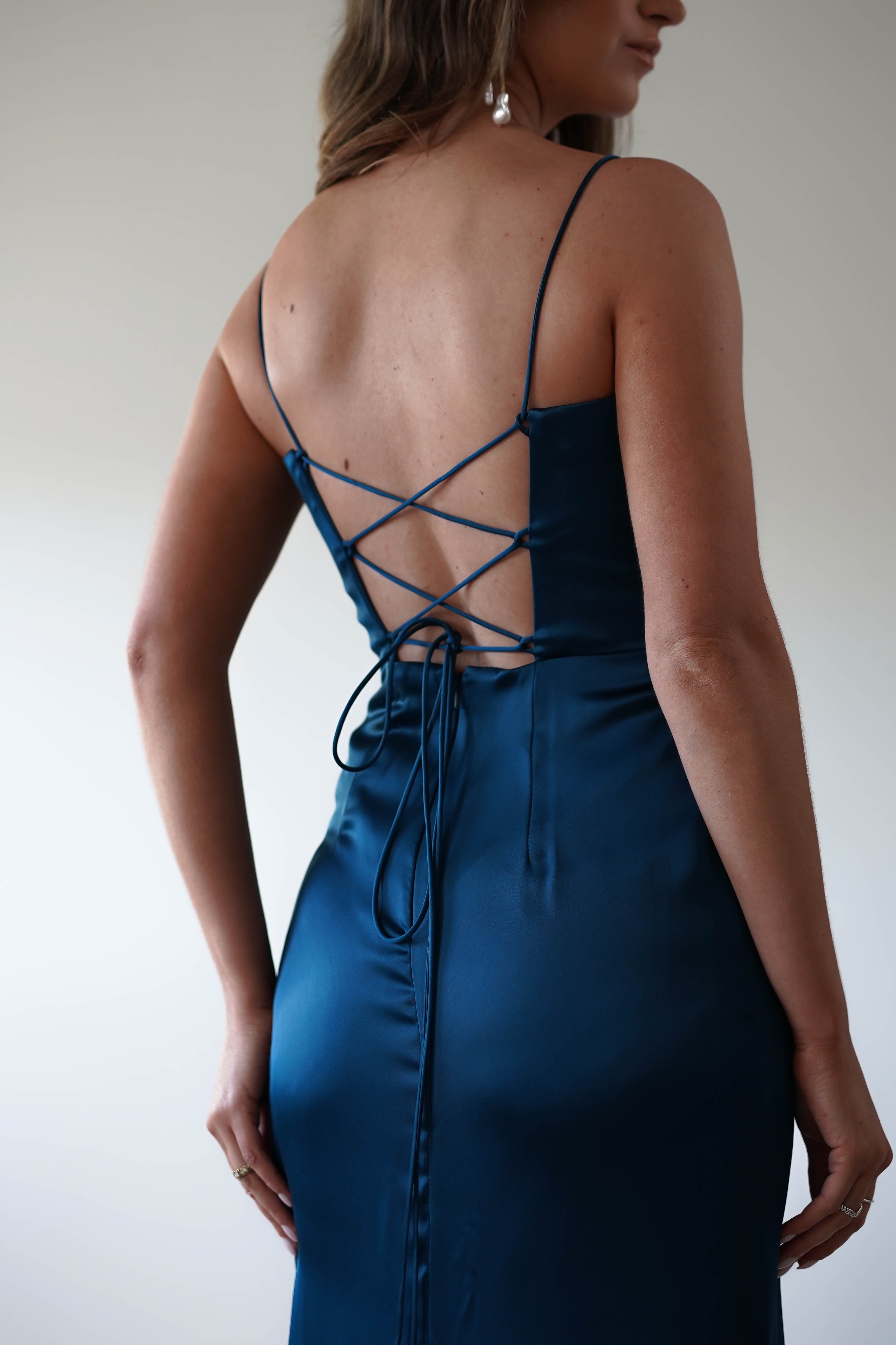 A person wearing a dark teal satin corset dress with thin straps and a laced open back design. The dress fits snugly and the scene is softly lit, highlighting its elegance—perfect for any special occasion.