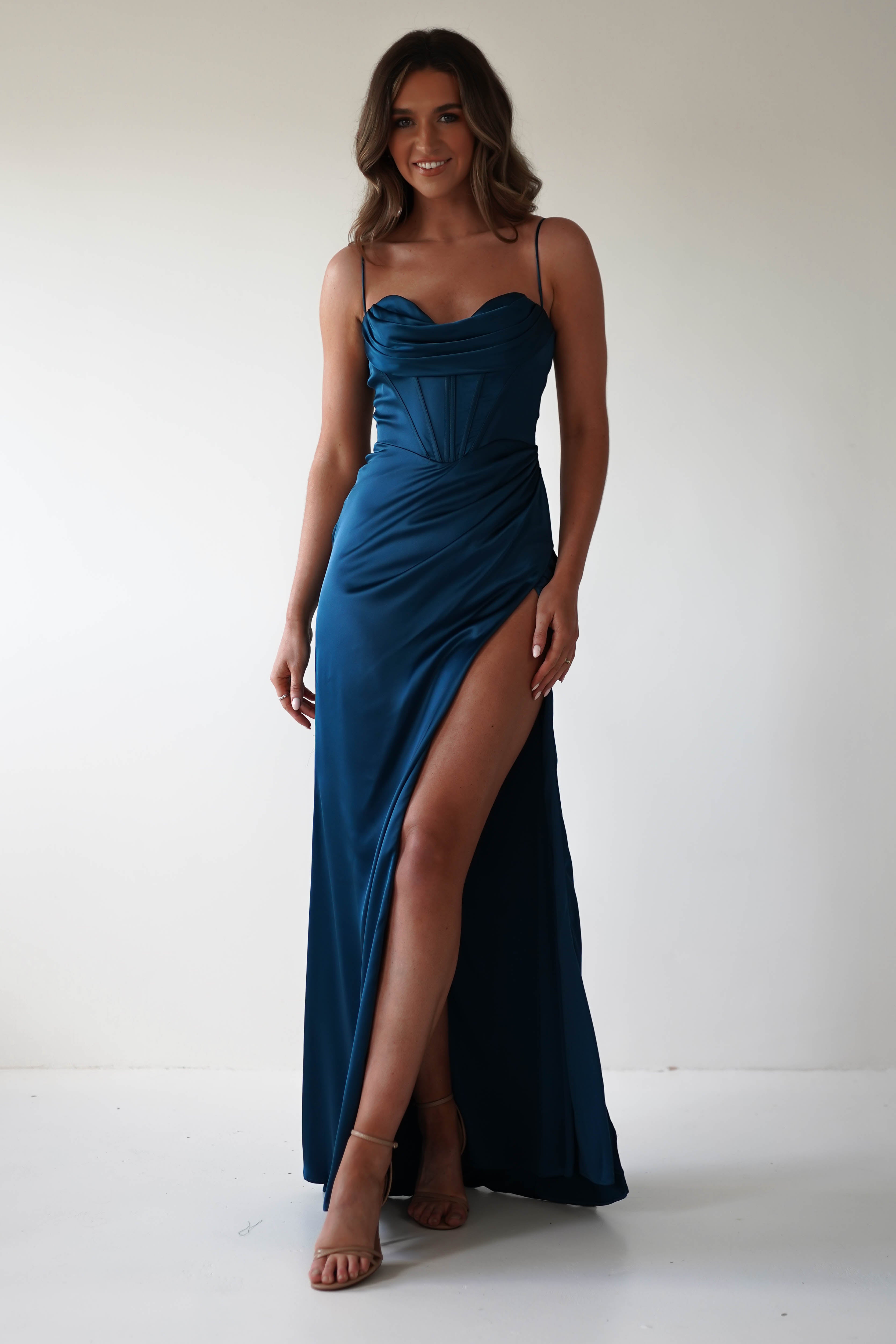 Woman in a teal satin gown with a thigh-high slit and spaghetti straps stands against a plain white background. Showcasing an adjustable fit for any special occasion, she pairs tan high heels with her look. Her long brown hair cascades as she smiles, one hand gracefully on her hip.