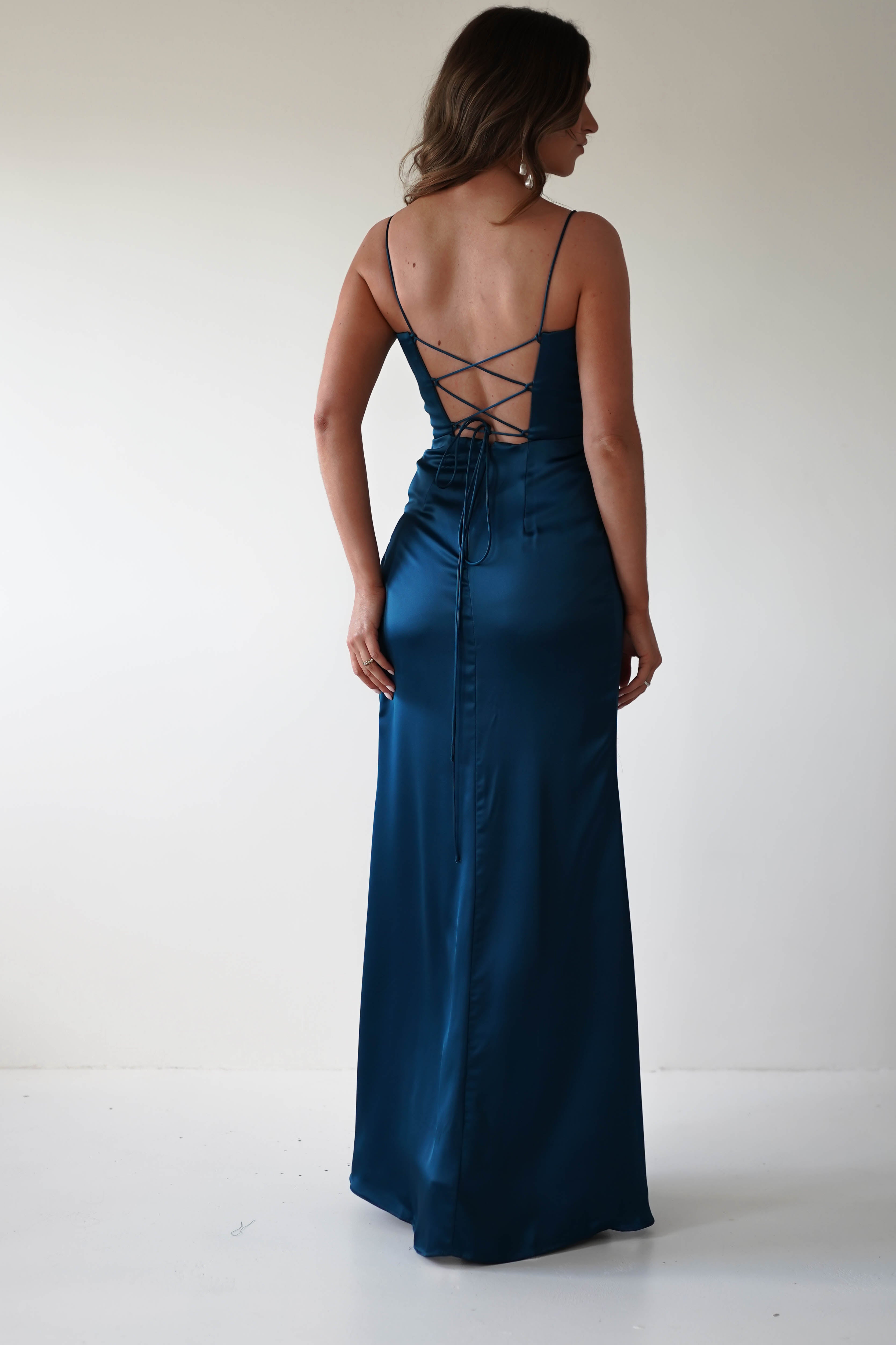 A woman stands facing away, wearing a teal satin corset dress with a lace-up back for an adjustable fit. Her wavy hair rests on her shoulders, perfectly complementing the floor-length gown with thin straps—a stunning choice for any special occasion against the plain white background.