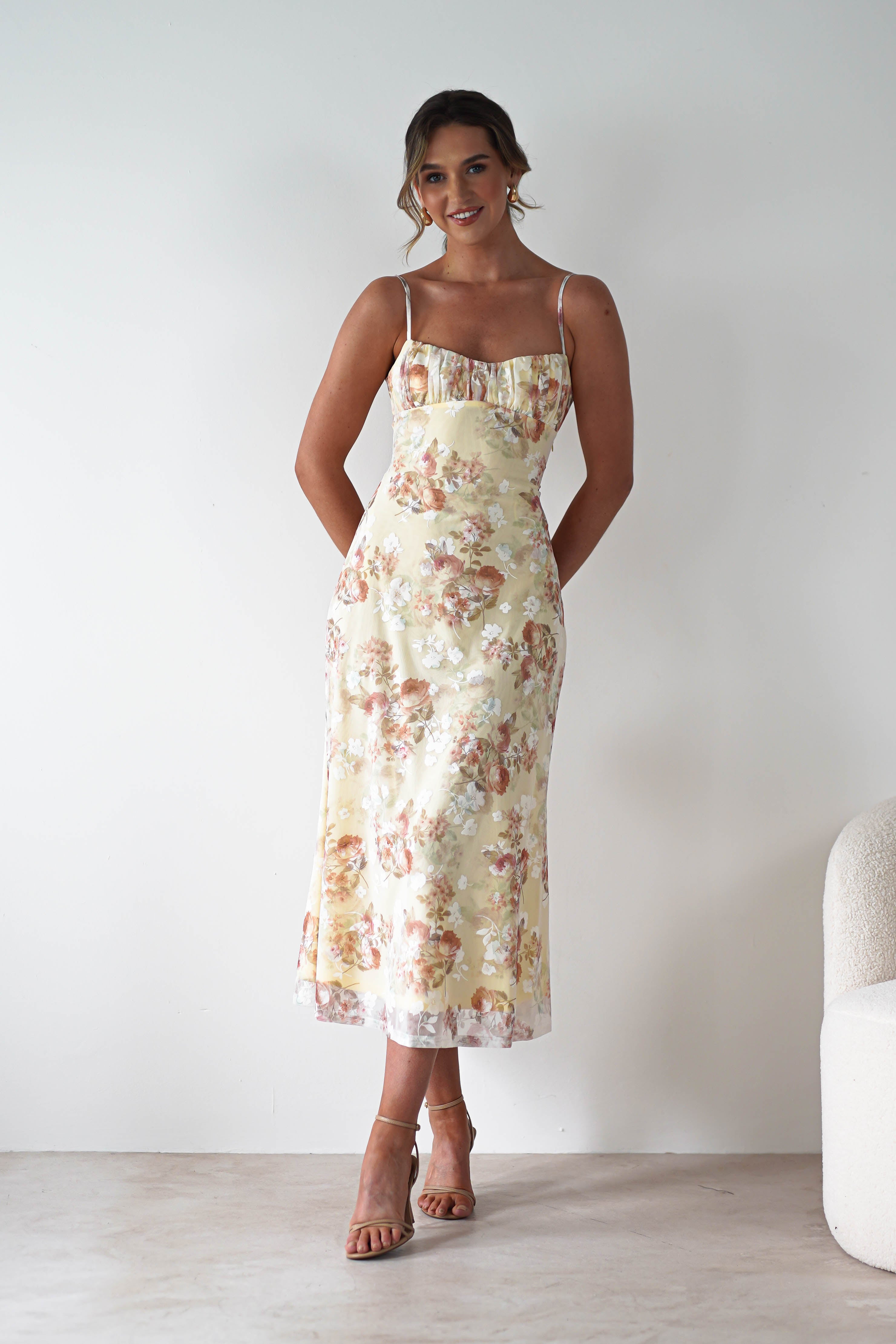 A woman stands against a plain white wall wearing a sleeveless, open back floral midaxi dress with thin straps and tan heeled sandals. She has her hands in her pockets and is smiling slightly—perfect for any wedding guest.