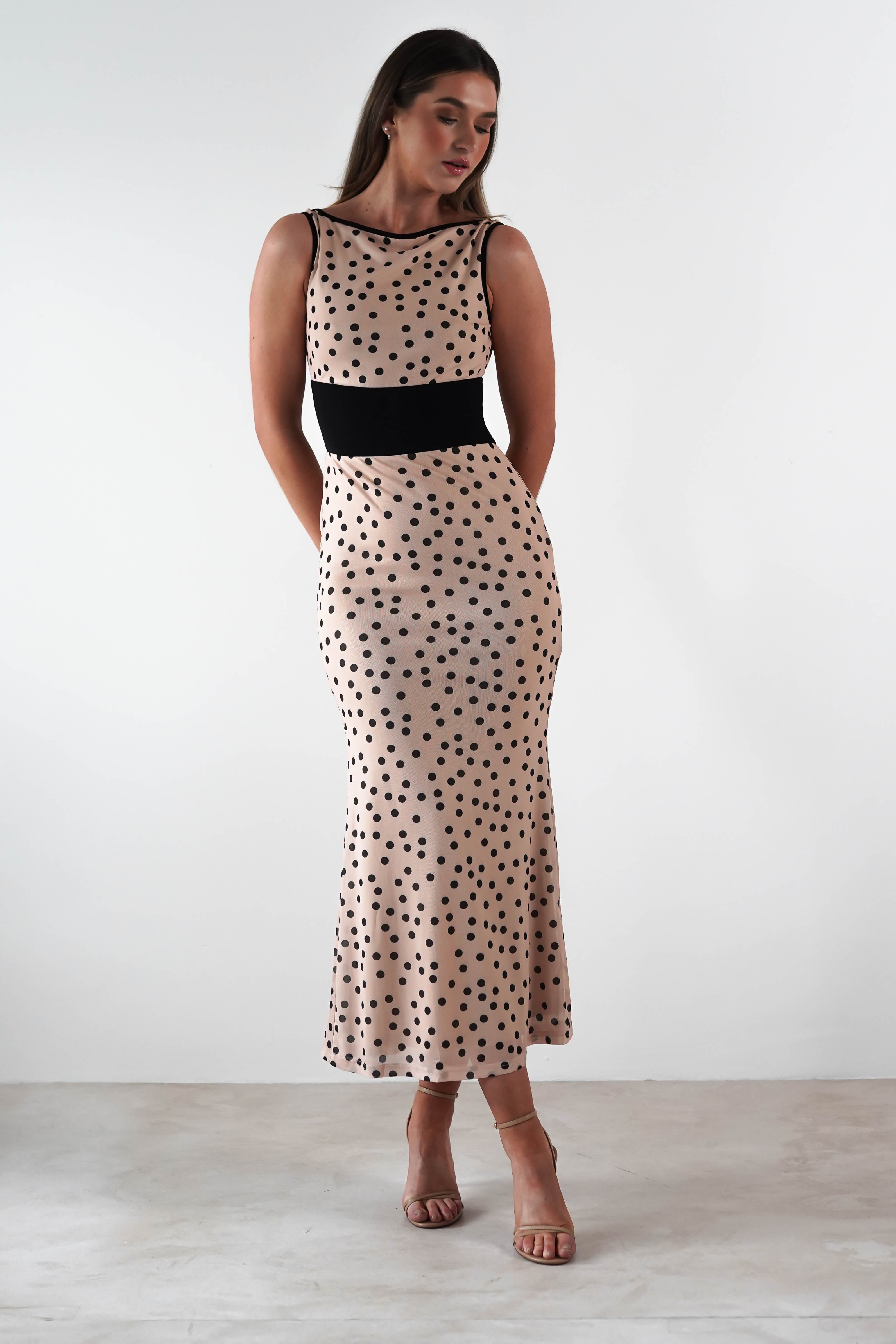 A woman is elegantly dressed in a sleeveless beige gown adorned with black polka print and a sleek waistband. Posed against a plain white backdrop, she gazes down with her hands in pockets, accentuating her strappy heels—a perfect ensemble for a wedding guest dress.