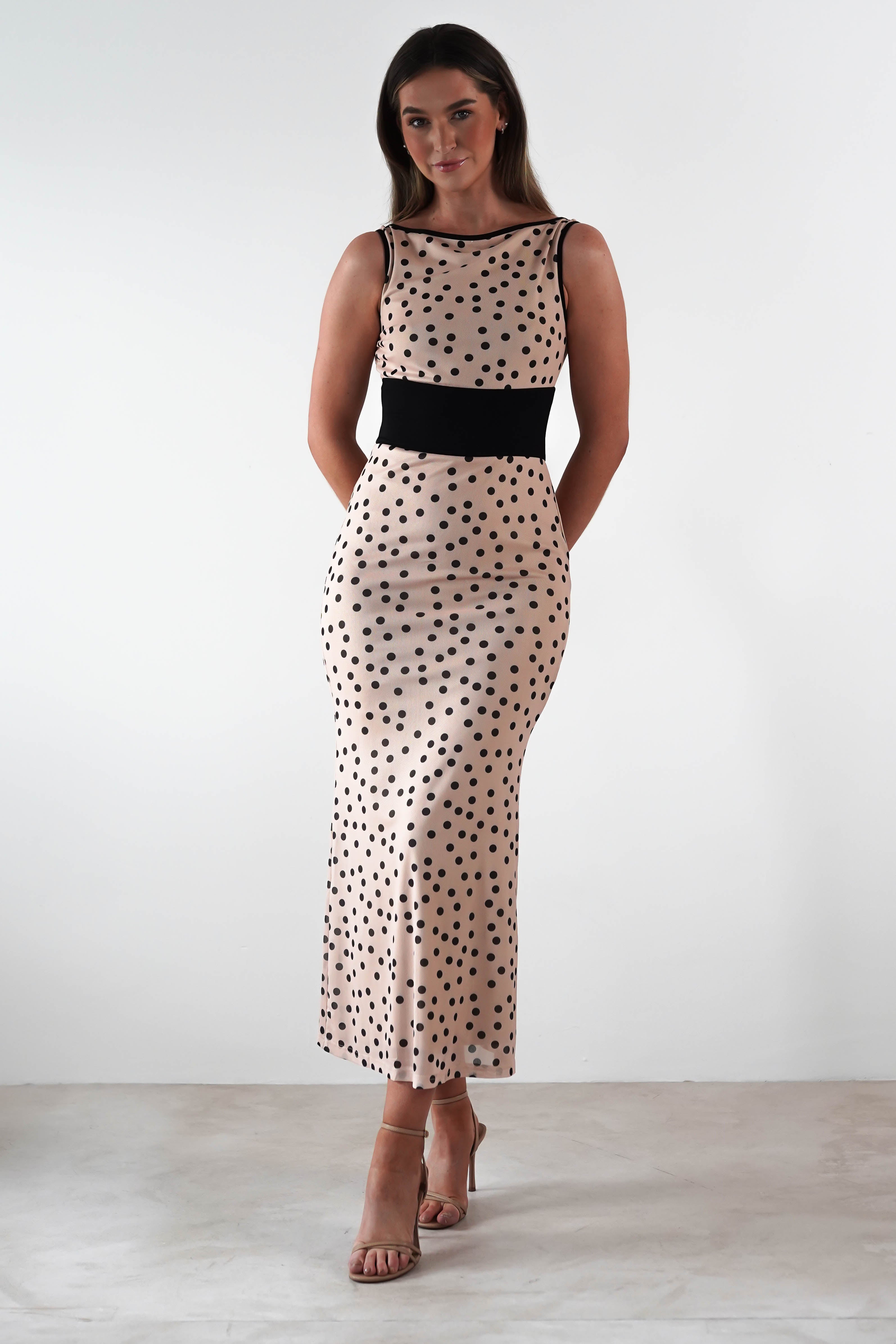 A woman stands against a plain white background, wearing a sleeveless, form-fitting beige wedding guest dress adorned with black polka dots and a black waistband. She has long hair and is wearing strappy high heels. Her hands are clasped behind her back.