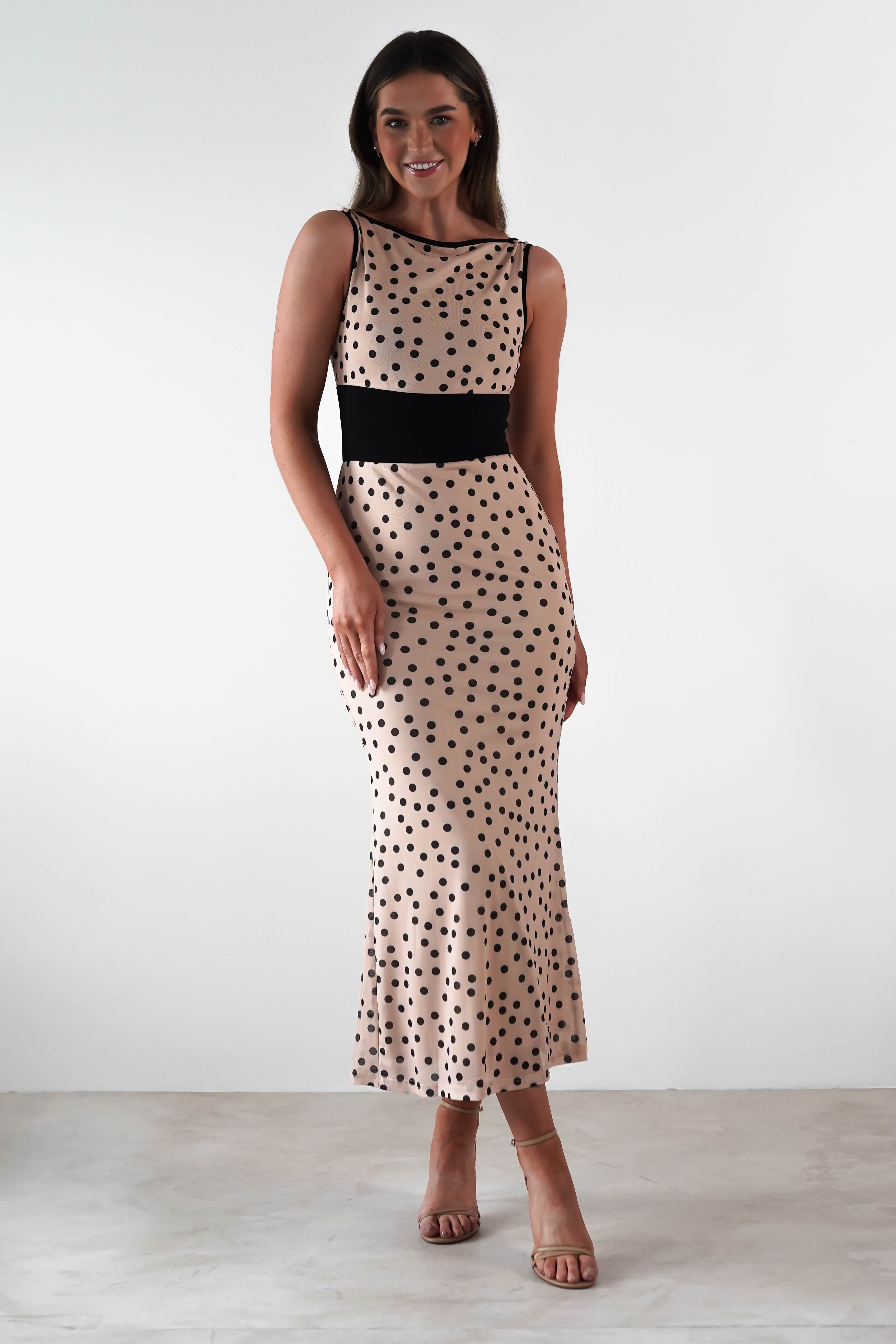 Woman in a sleeveless, beige wedding guest dress with black polka dots and a black waistband stands against a plain white background. She has long dark hair and is wearing strappy heels, looking at the camera with a slight smile.