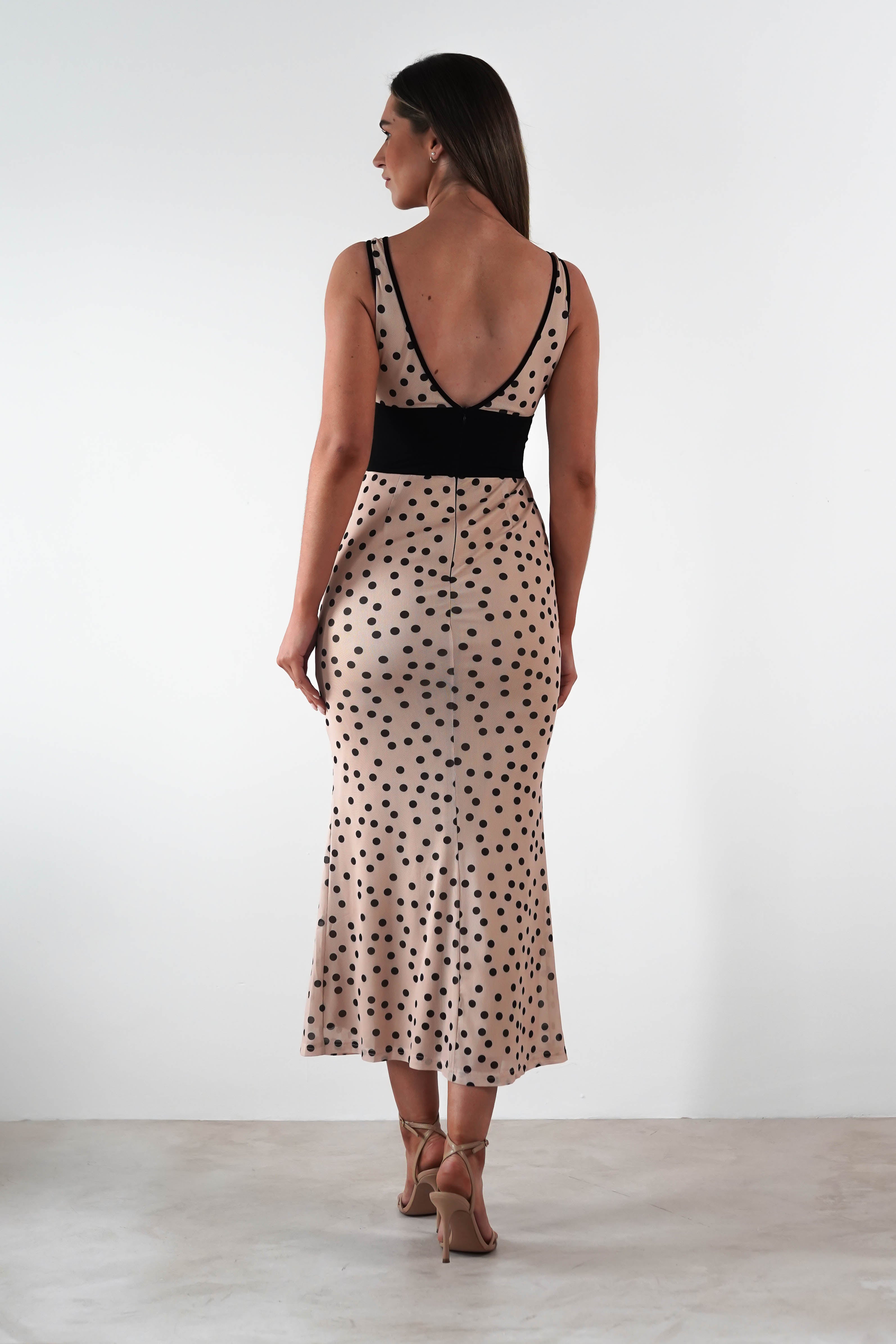 A woman in a sleeveless beige midi dress with black polka dots and a deep V back stands on a light-colored floor, exuding the elegance of a wedding guest. She wears tan high heels and has long, straight brown hair against the plain white background.