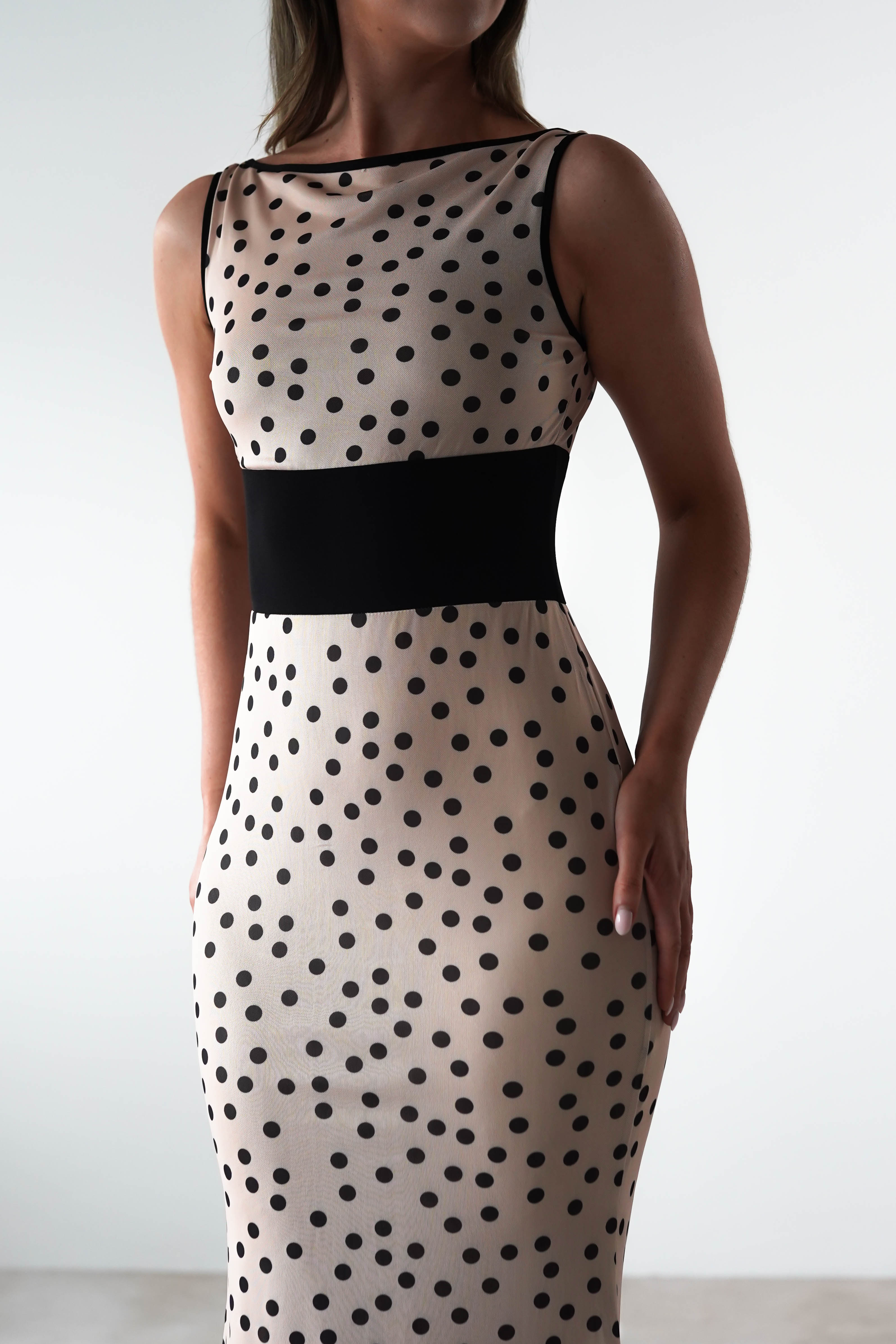 A person stands against a plain background wearing a sleeveless, polka print dress with black dots. The dress features a square neck and is complemented by a wide black waistband, making it an ideal choice for a wedding guest.