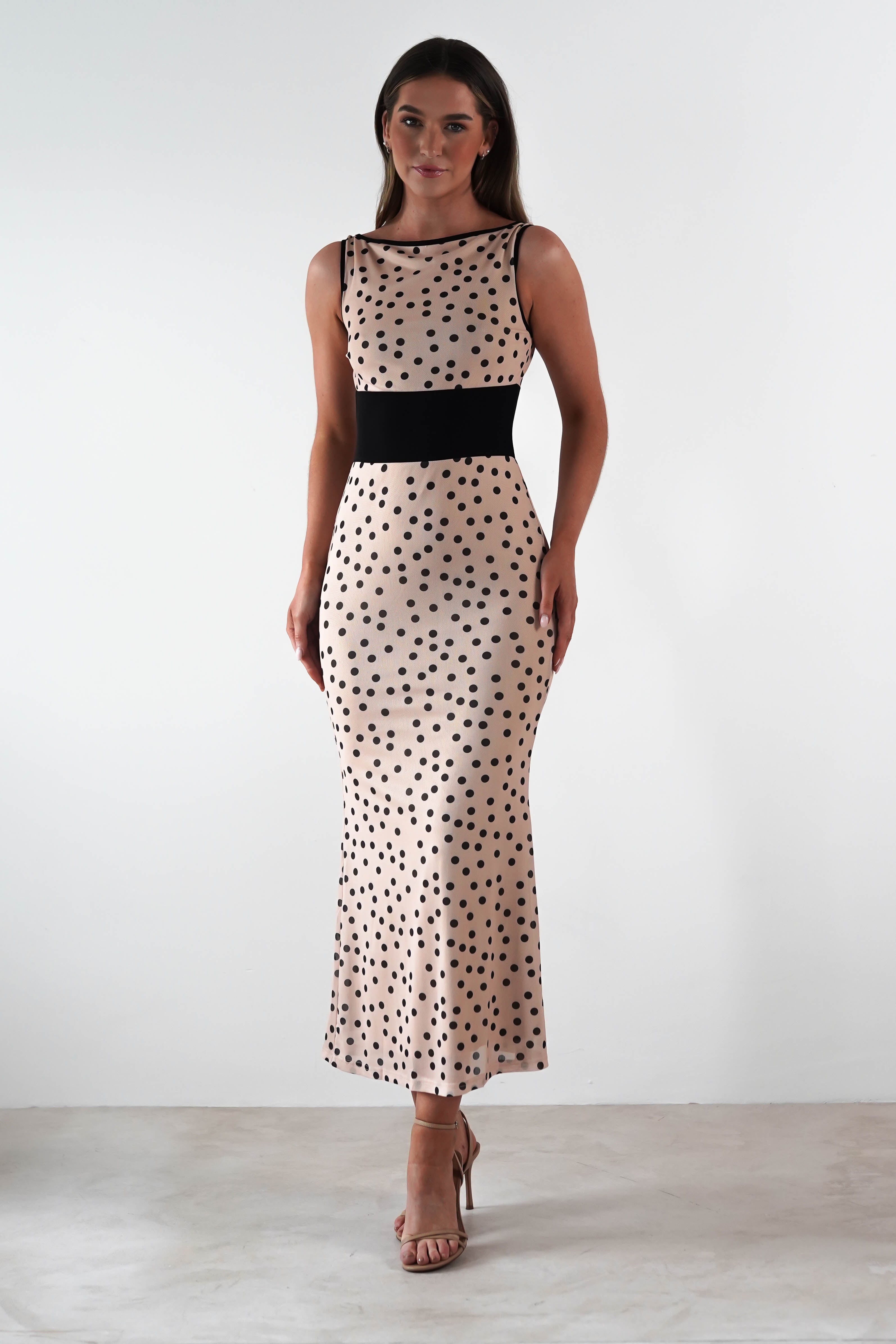 Person in a sleeveless, fitted beige dress with black polka dots and a square neck, perfectly suited as a stylish wedding guest dress. They are standing on a light-colored floor, wearing open-toed heels, with a neutral expression against the plain white background.