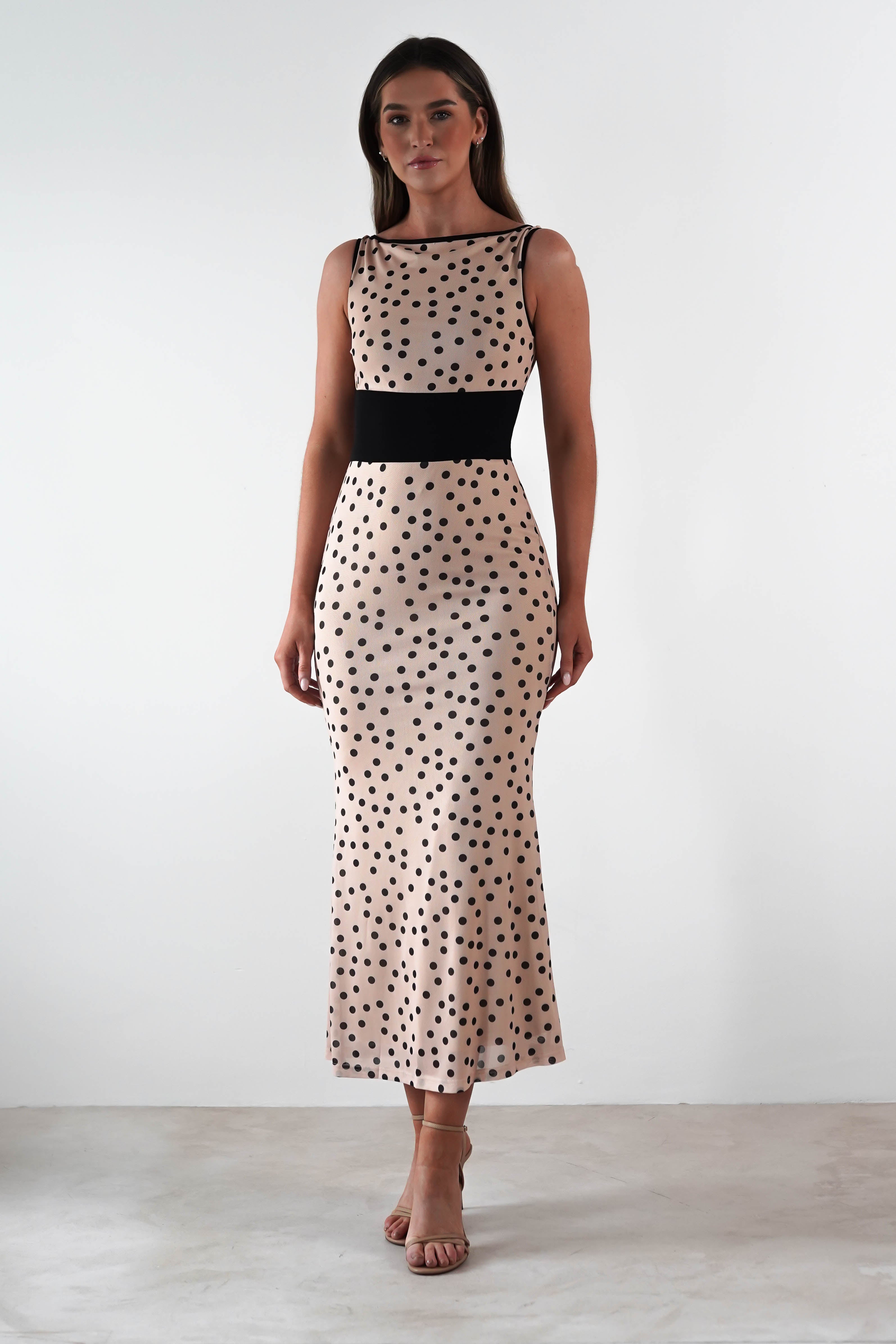 A person is elegantly attired in a sleeveless square neck maxi dress, adorned with black polka dots and a chic black waistband. Paired with strappy beige heels, this ensemble perfectly suits any wedding guest attire, set against a plain white backdrop.