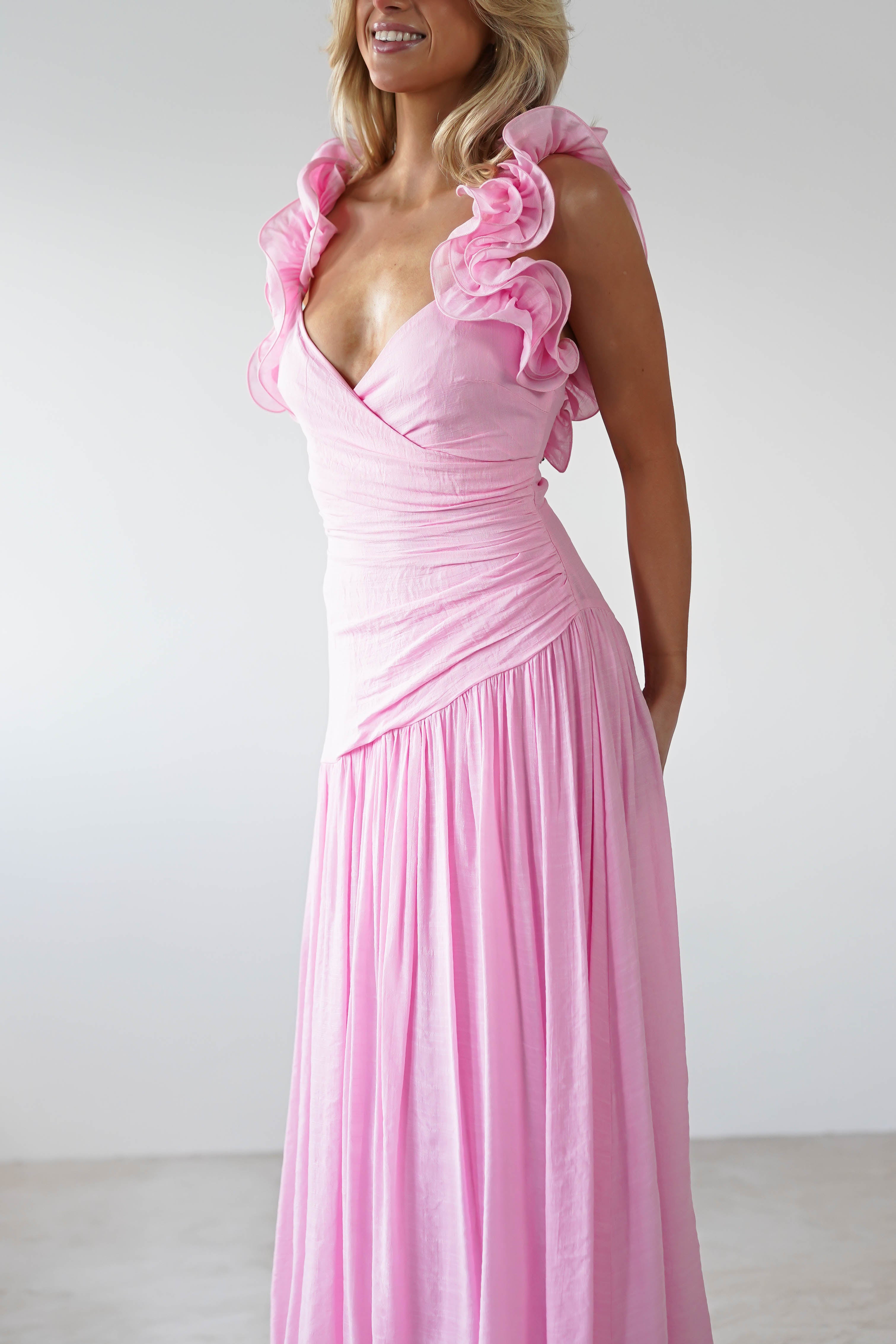 A person is wearing a flowing pink midaxi dress with elegant ruffle shoulders, perfect as a wedding guest outfit. The dress features a fitted bodice and a long, pleated skirt, set against a plain white background.