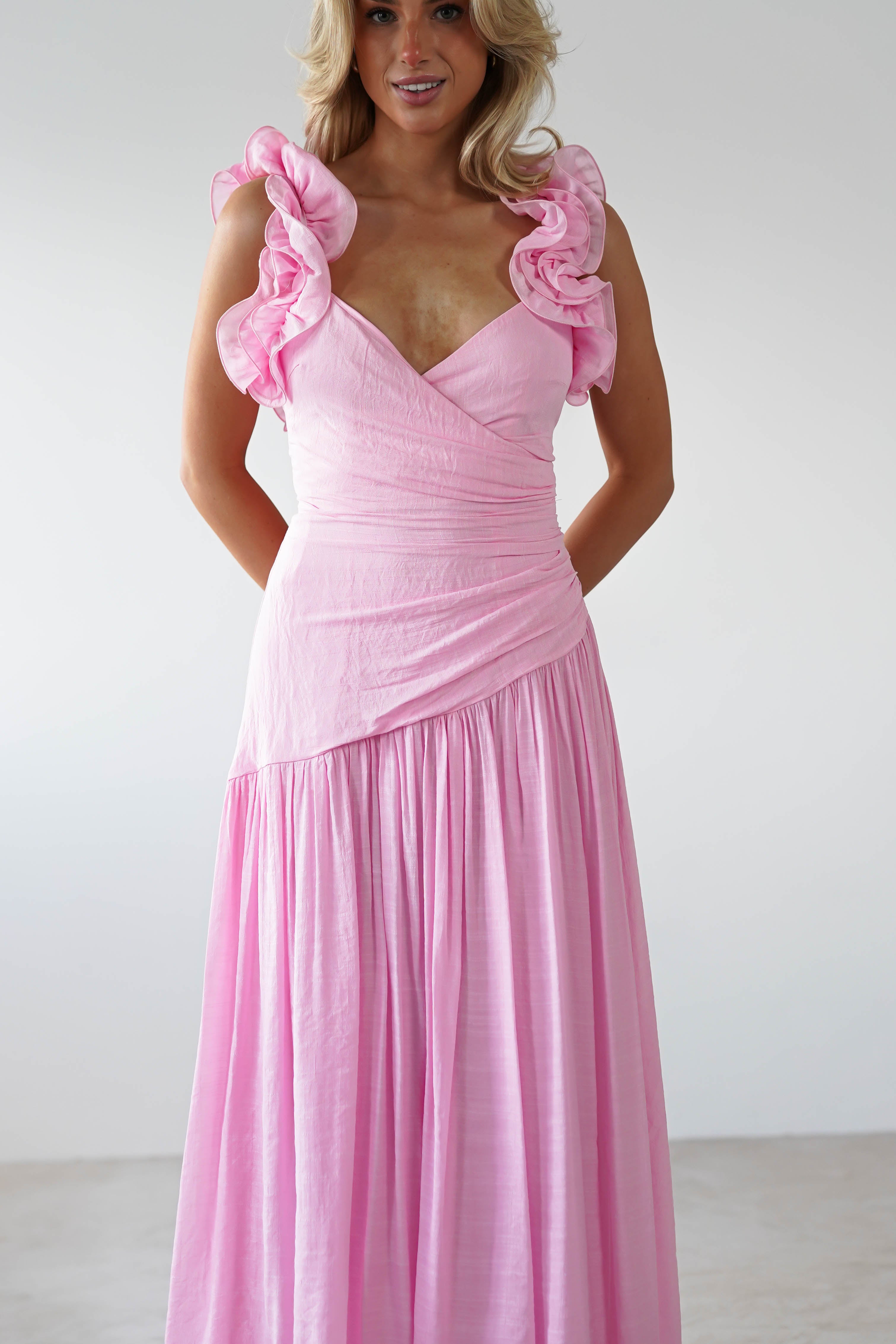 A woman stands gracefully in a pink midaxi dress with elegant ruffle shoulders, its flowing skirt draping beautifully against the plain background. Perfect as a wedding guest outfit, this ensemble exudes charm and sophistication.