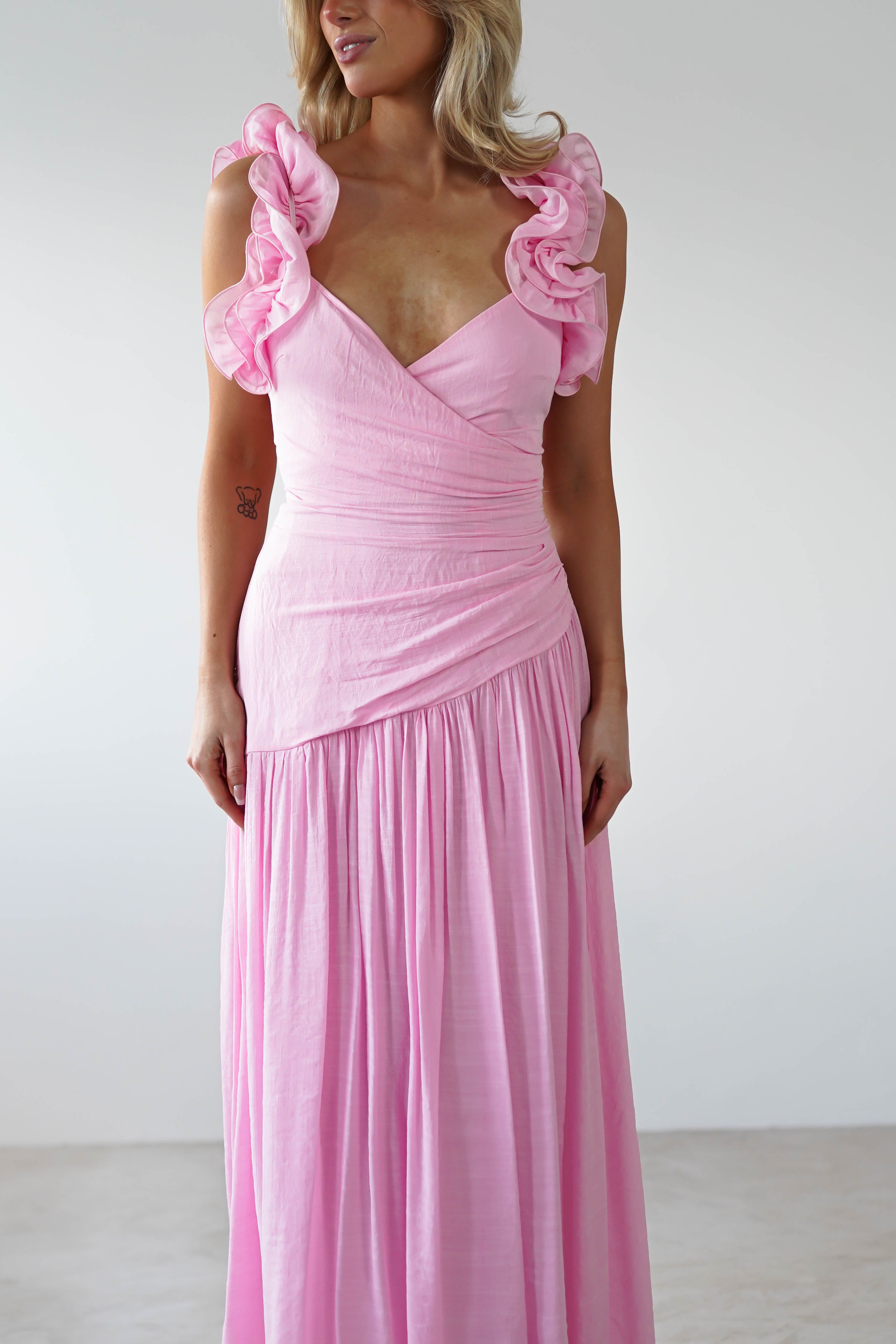 A person wearing a stunning pink midaxi dress with ruffle shoulders and a wrap-style bodice. The elegant wedding guest outfit features a flowing, gathered skirt against a plain white background.