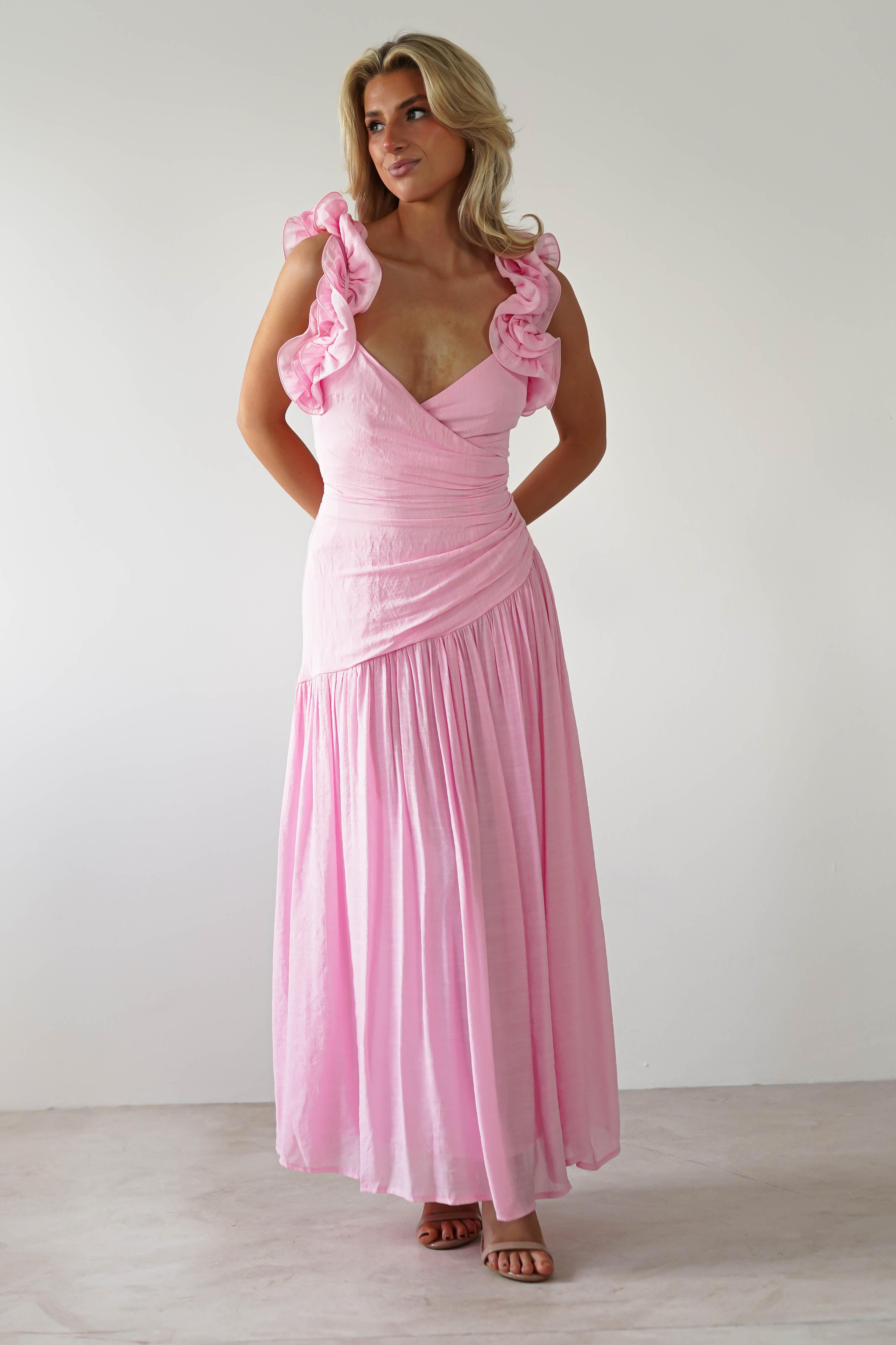 A woman in a pink midaxi dress with ruffle shoulders stands against a plain background. Her hands are behind her back, and she gazes slightly to the side. The elegant ensemble subtly suggests its an ideal wedding guest outfit.