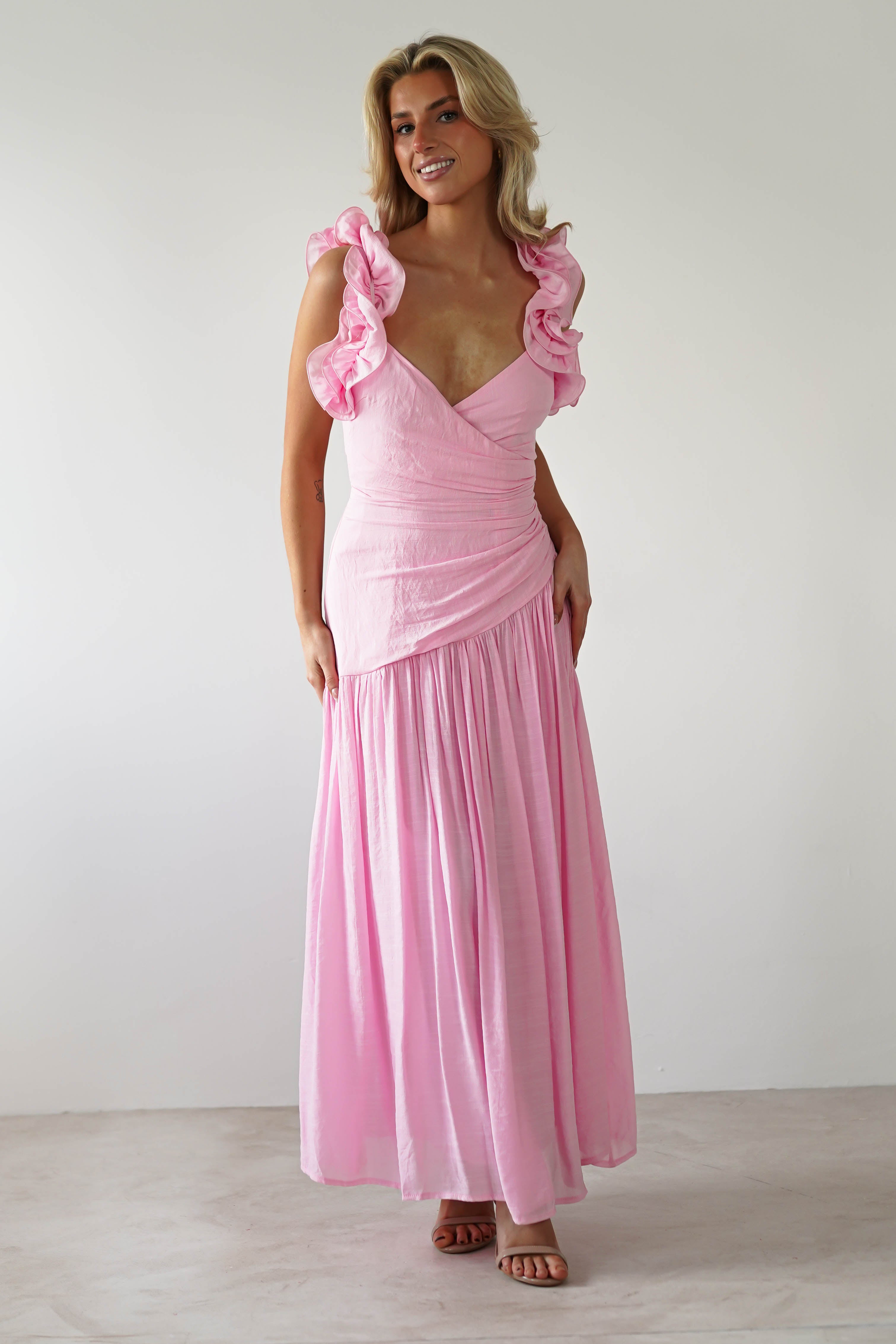 A smiling person stands gracefully in a pink midaxi dress with ruffle shoulders, set against a plain white background.