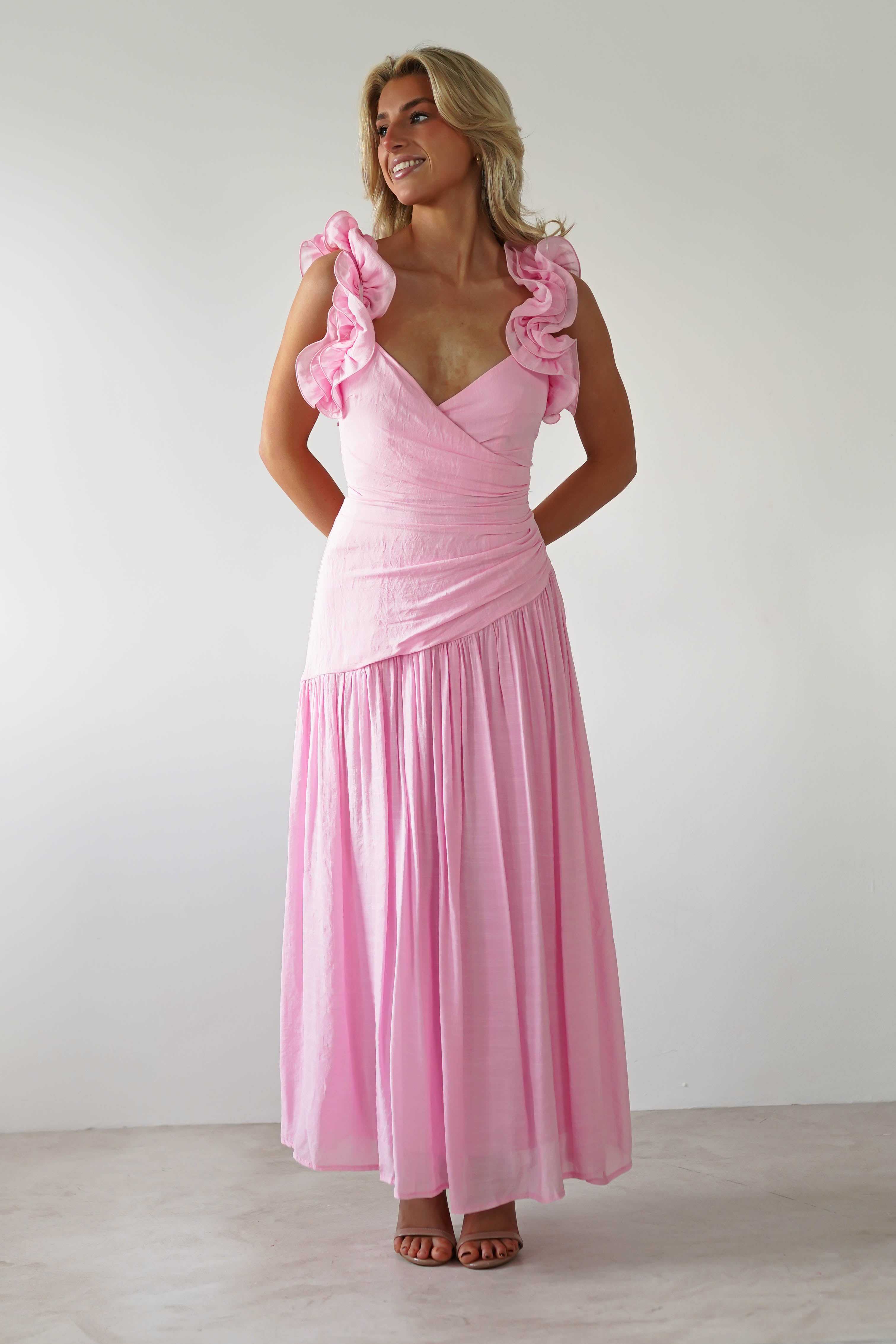 A person wearing a long pink midaxi dress with ruffled shoulder straps stands against a plain white background. They are looking to the side, hands clasped behind their back, and smiling—a perfect choice for wedding guest dresses.